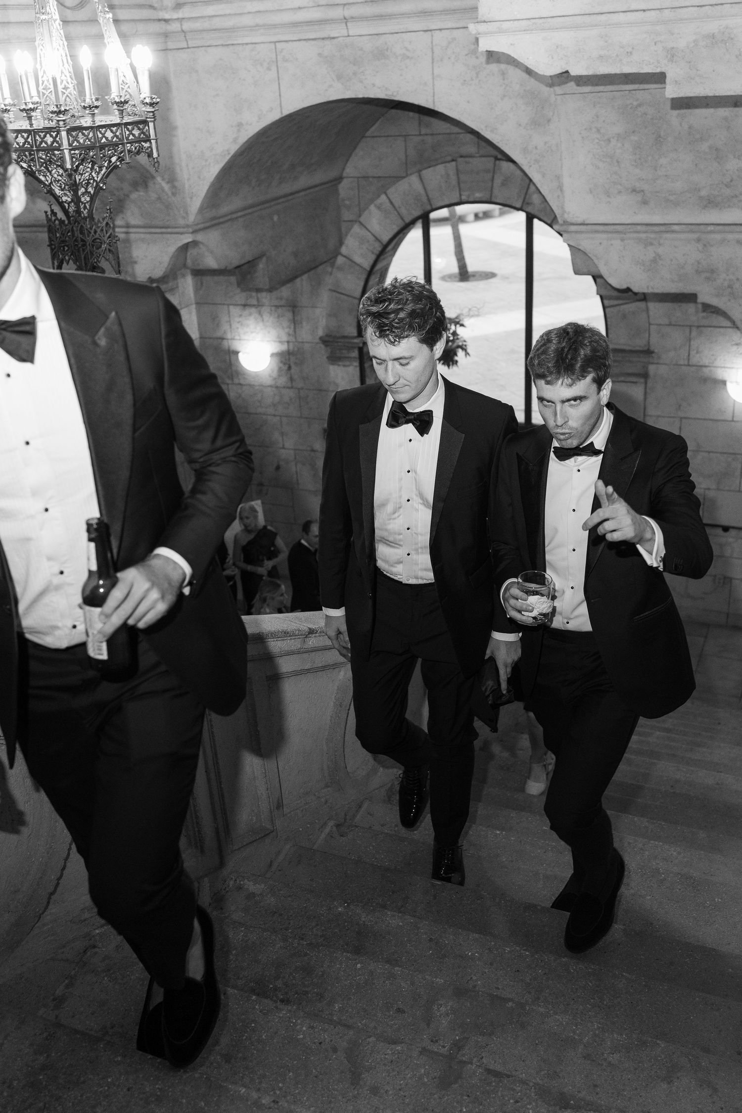 Three men in black tuxedos descend grand stone staircase at formal event in black and white photo.