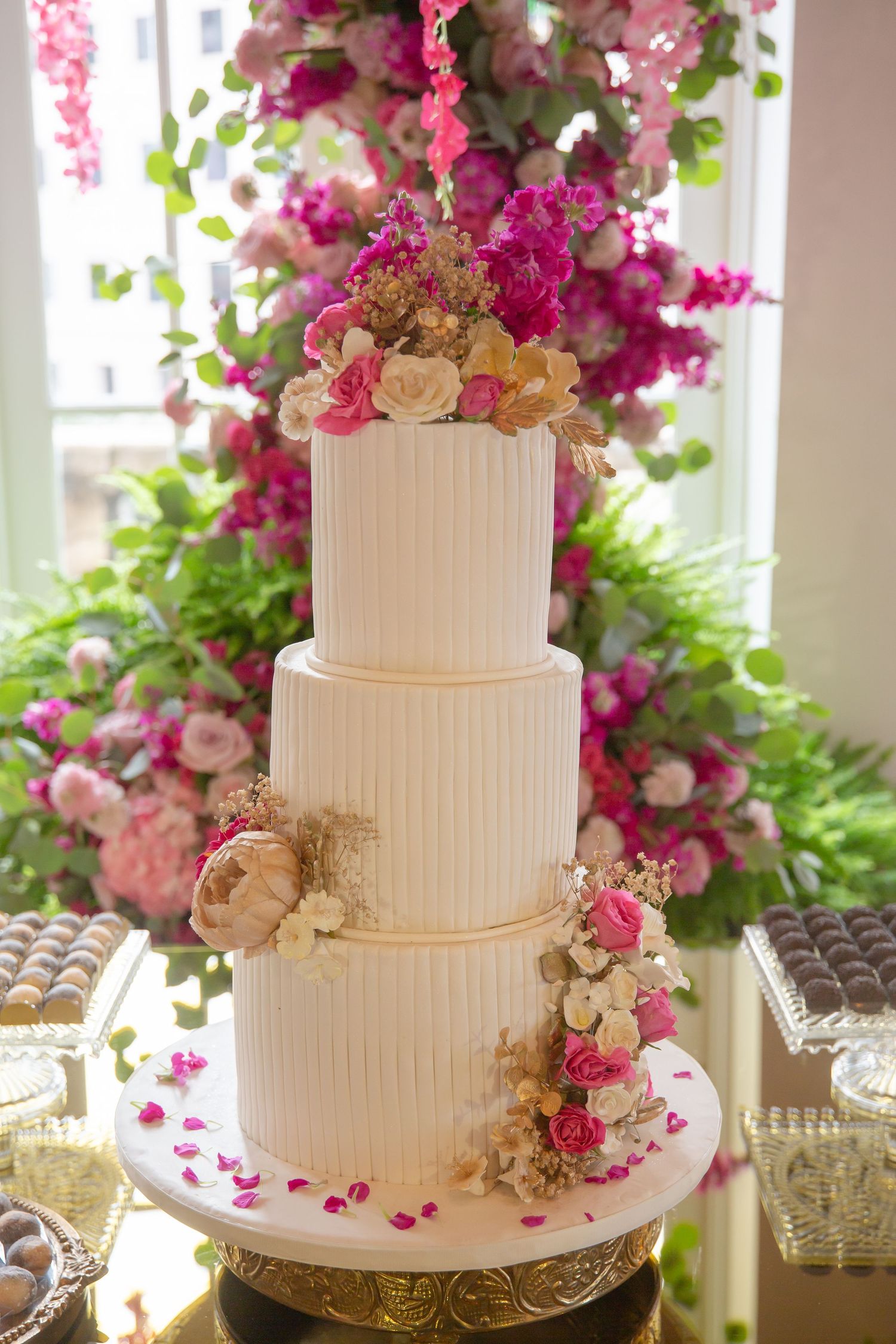 Elegant three-tier white wedding cake adorned with pink and cream flowers, displayed against a lush floral backdrop.