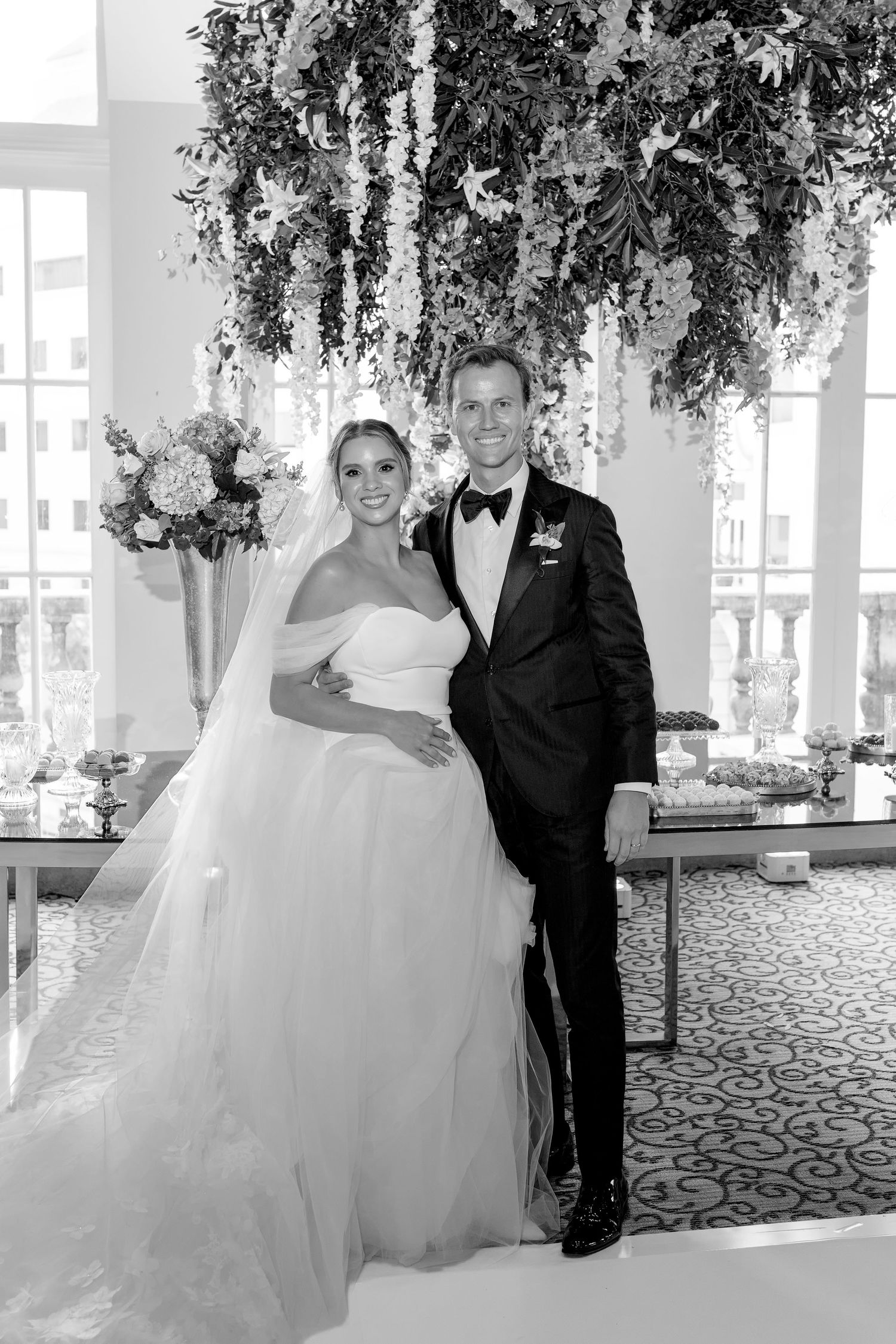 Black and white wedding photo of bride in strapless gown and groom in tuxedo posing under floral arrangement.