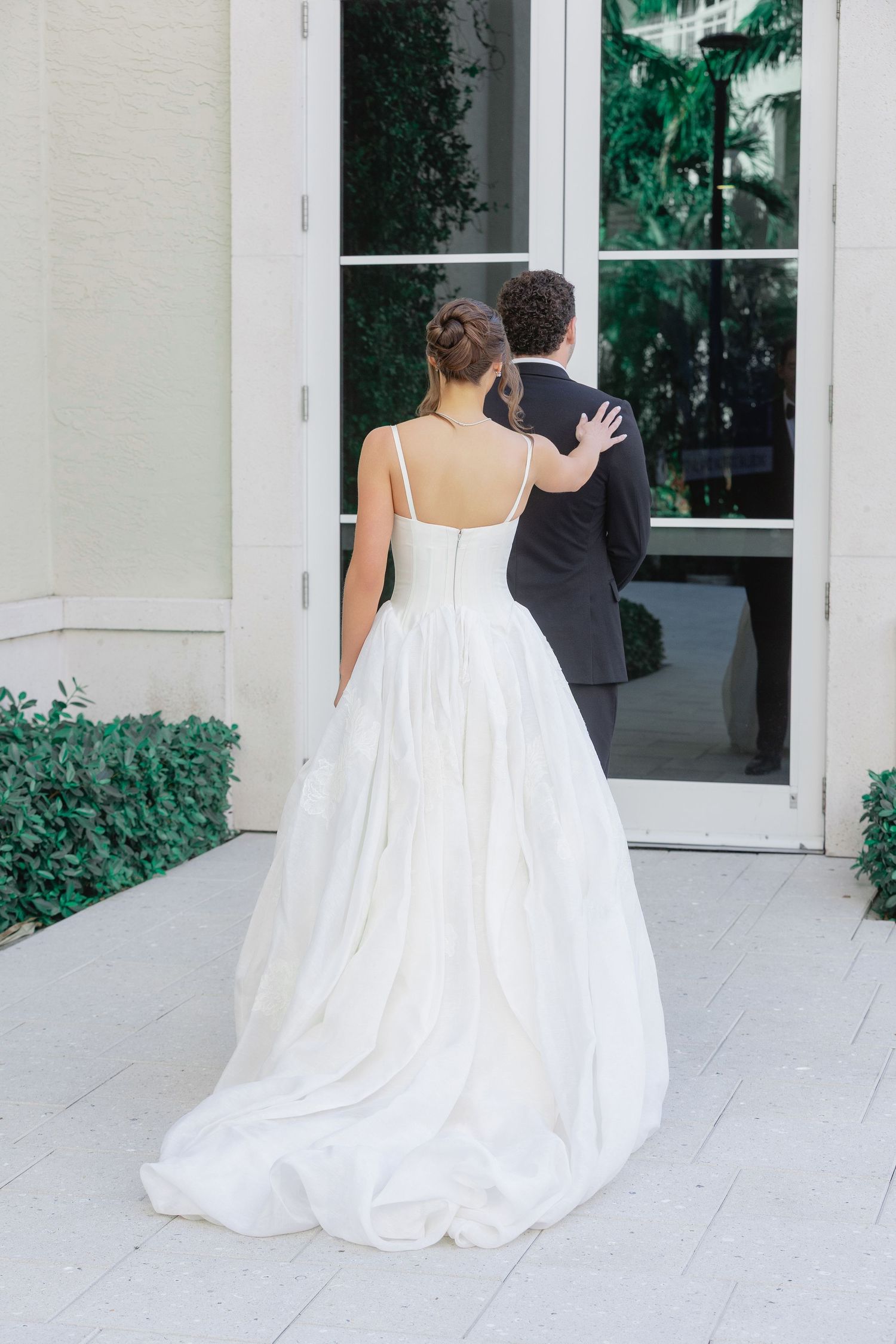 Bride in white ballgown with spaghetti straps stands with groom in black tuxedo outside elegant venue doors.
