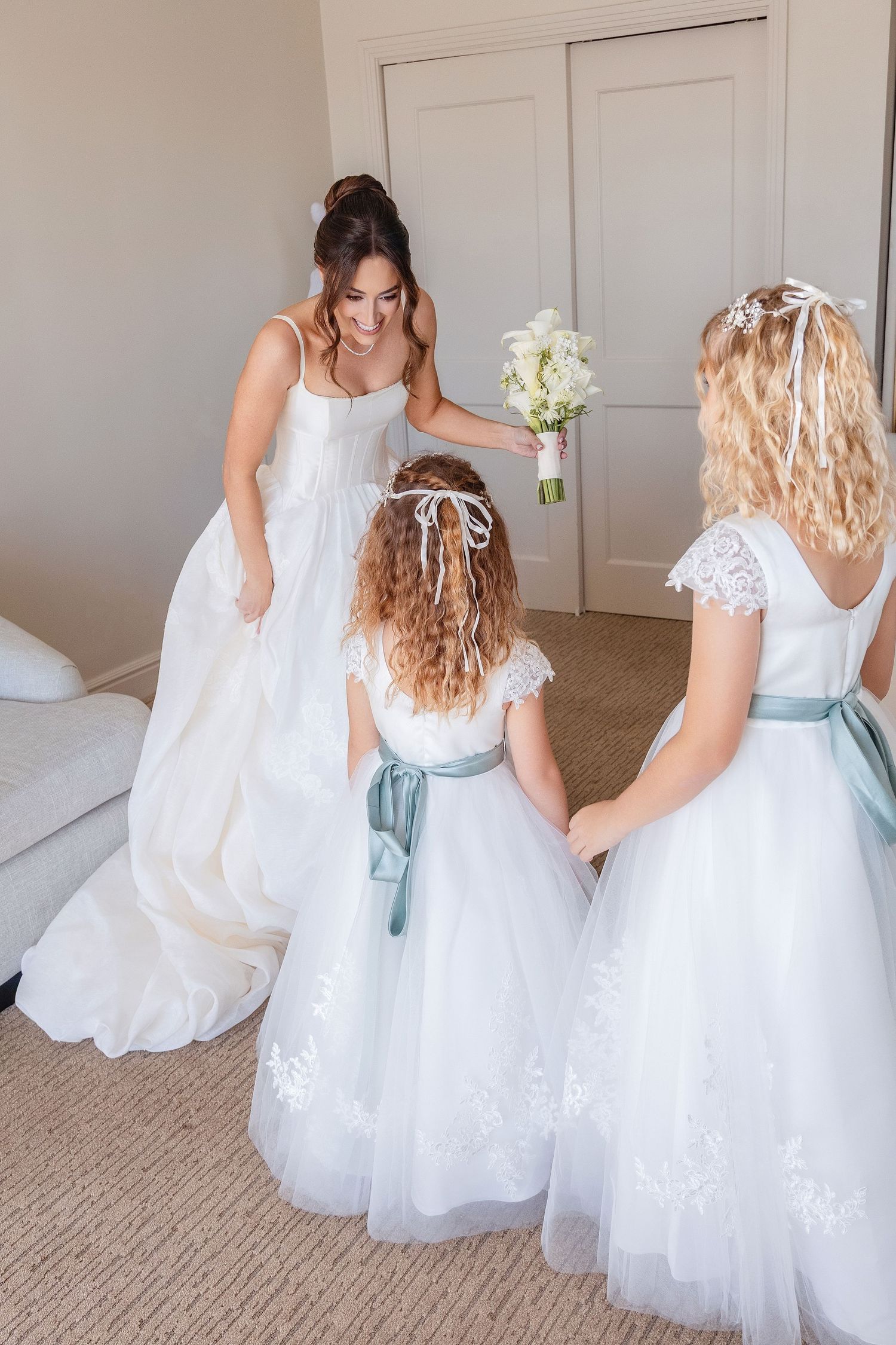 Bride in white gown holds bouquet while two flower girls in white dresses with sage green sashes stand nearby indoors.