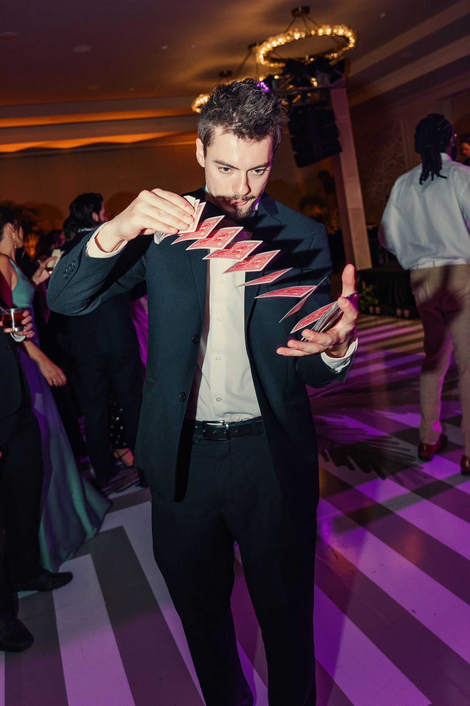 A magician in a black suit performs a card trick on a dance floor at an elegant evening event.