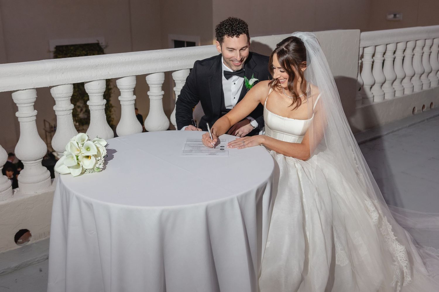 Bride and groom signing wedding documents at a white draped table, smiling together in elegant formal attire.
