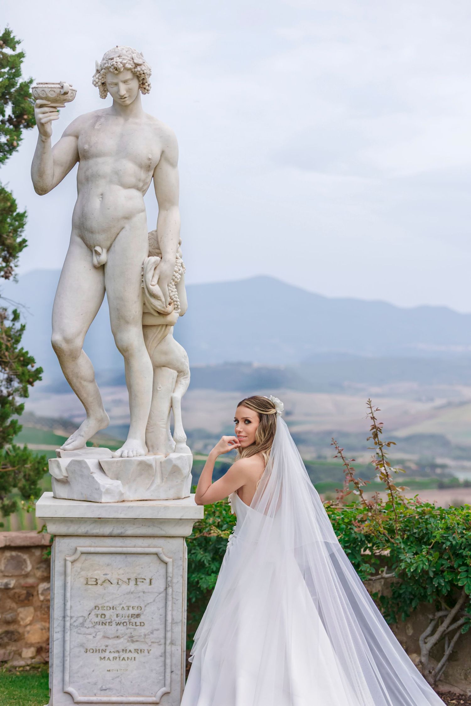 Classical white marble statue stands beside flowing wedding dress in Italian garden setting.