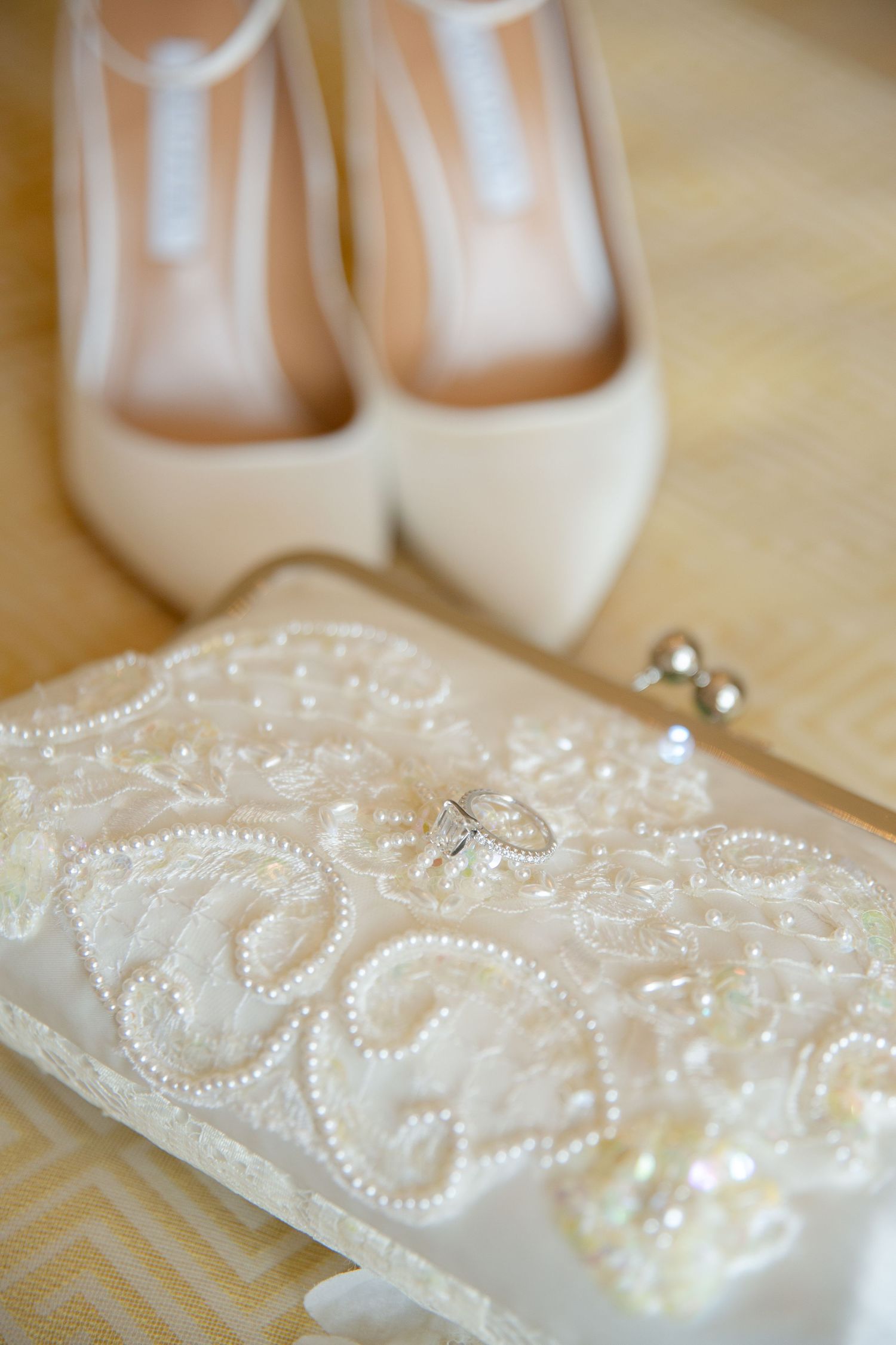 Elegant bridal accessories featuring white lace clutch purse, diamond ring, and ivory pointed-toe heels on wooden floor.