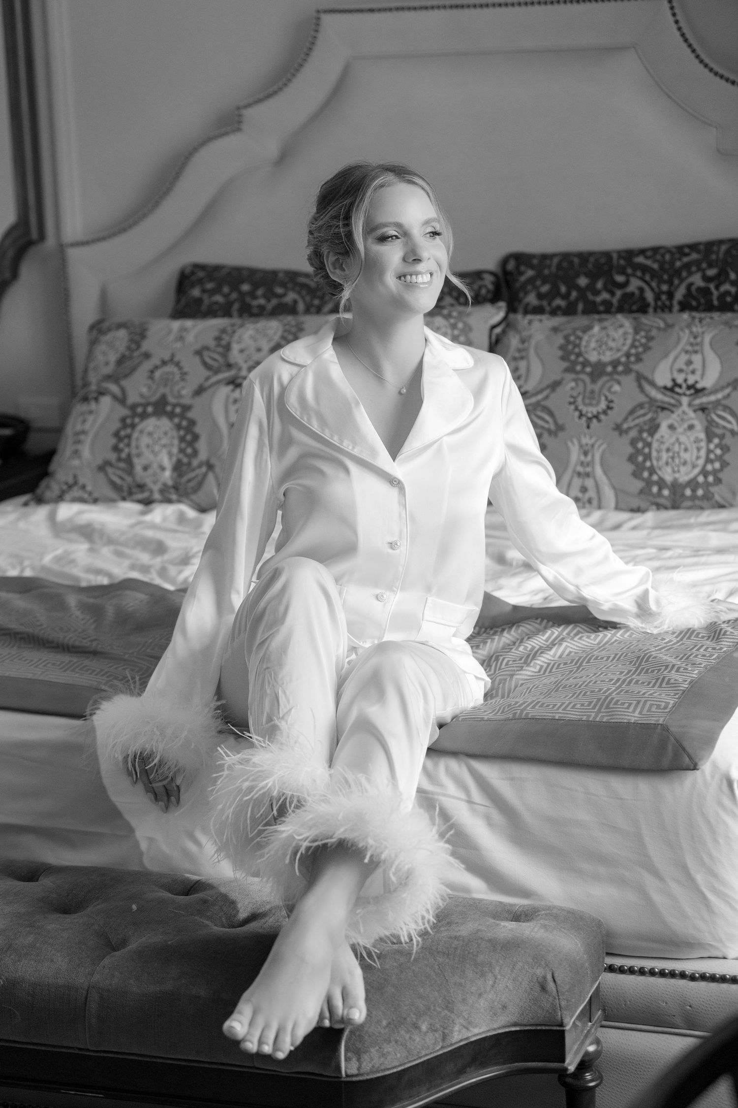 Smiling bride in white satin feather-trimmed pajamas sitting barefoot on bed in black and white photo.