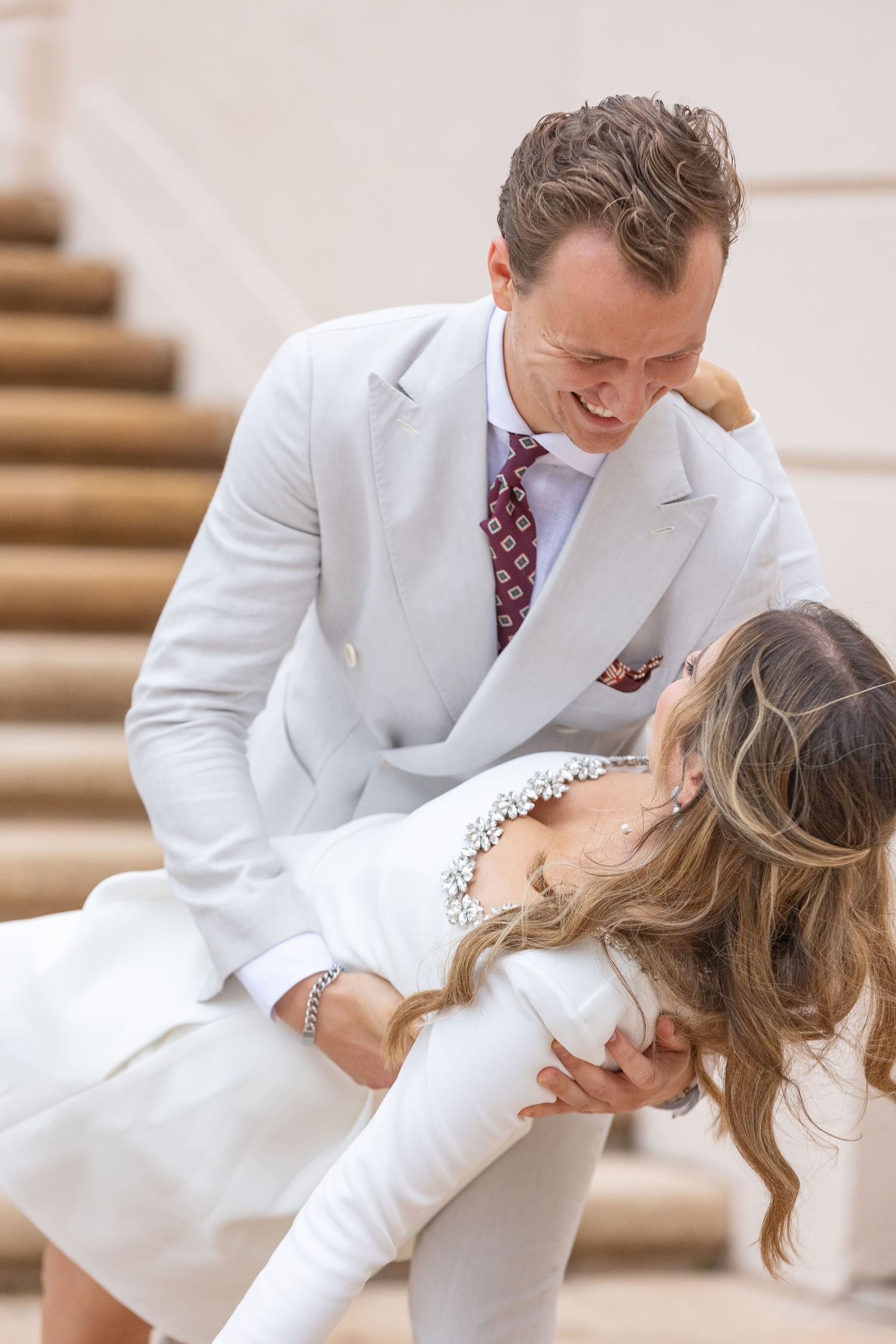 Couple in white outfits laughing together on outdoor steps, man in light suit with burgundy tie dipping woman.