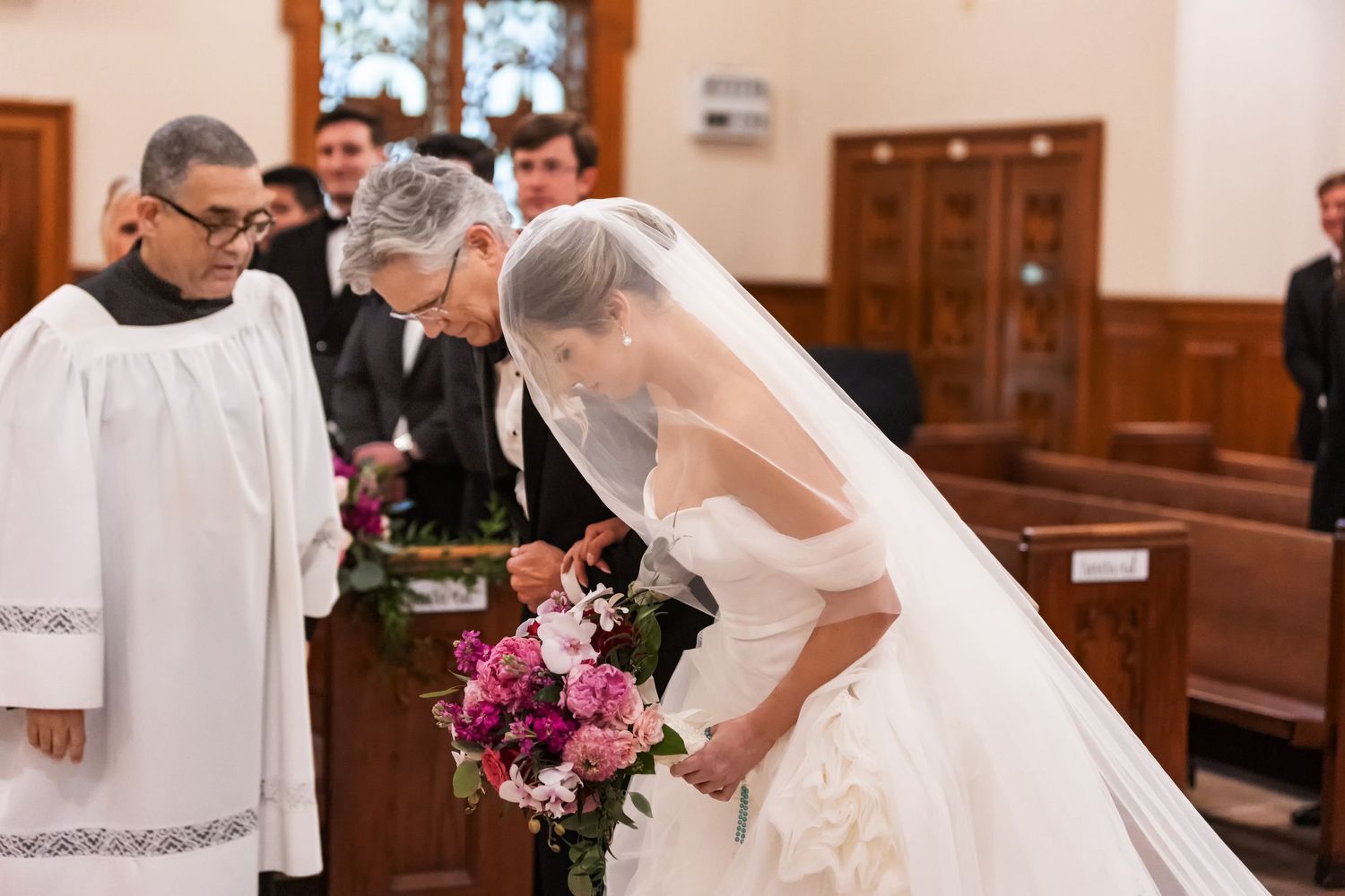 Bride in white gown and veil bowing with bouquet before priest in church ceremony.