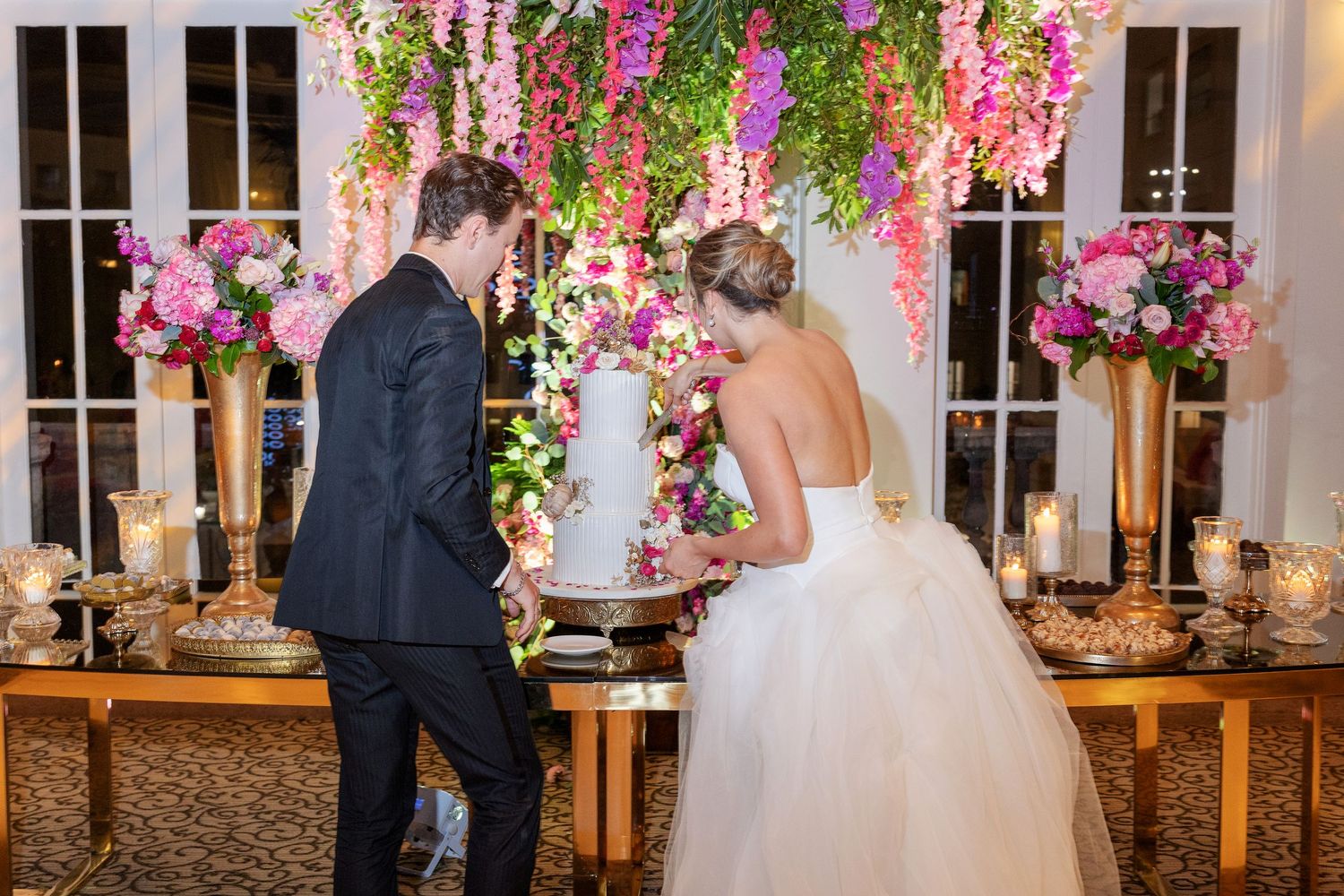 Bride and groom cutting wedding cake beneath lush pink floral arch at elegant evening reception.