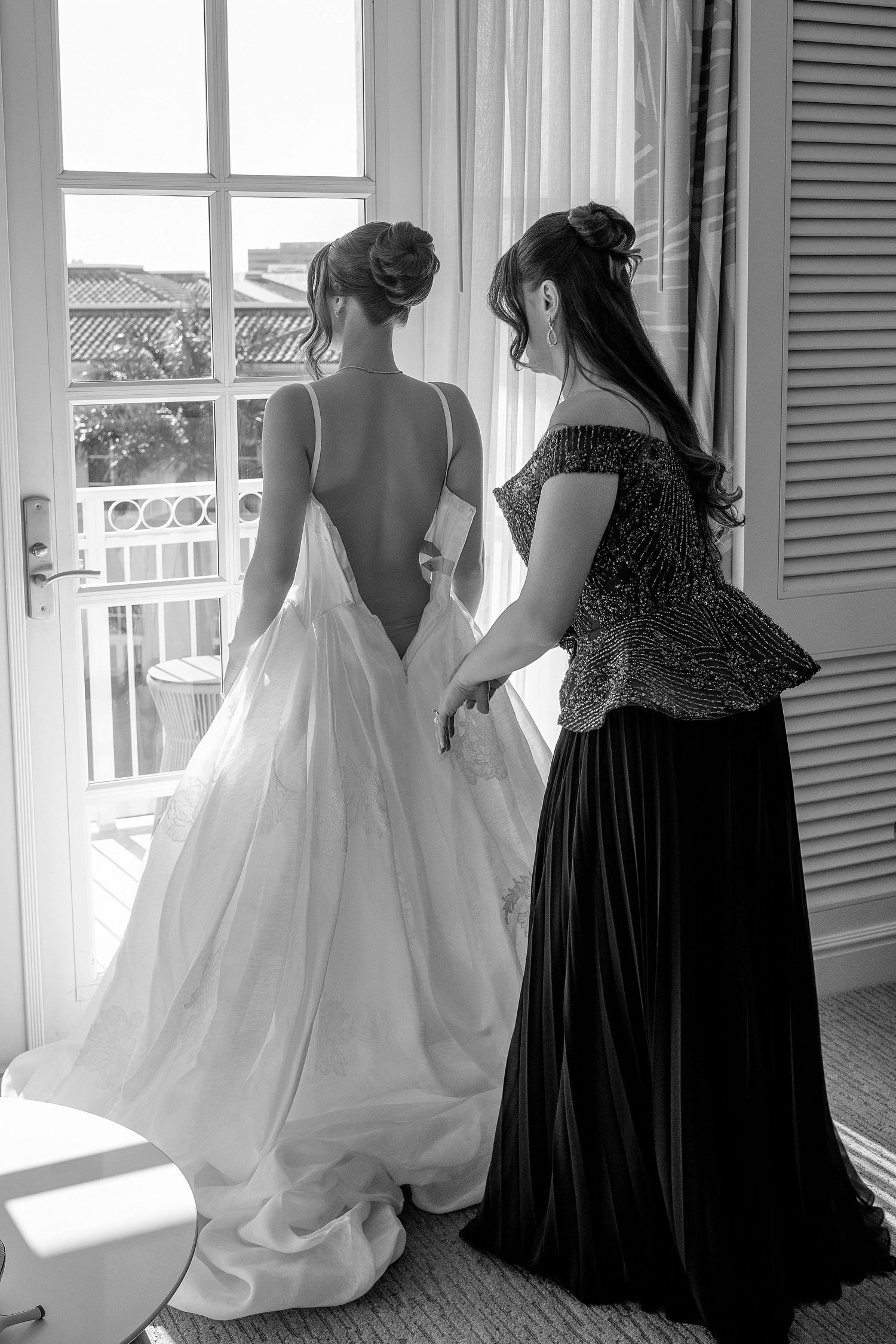 Black and white photo of bride in backless wedding gown being assisted by bridesmaid near sunlit French doors.