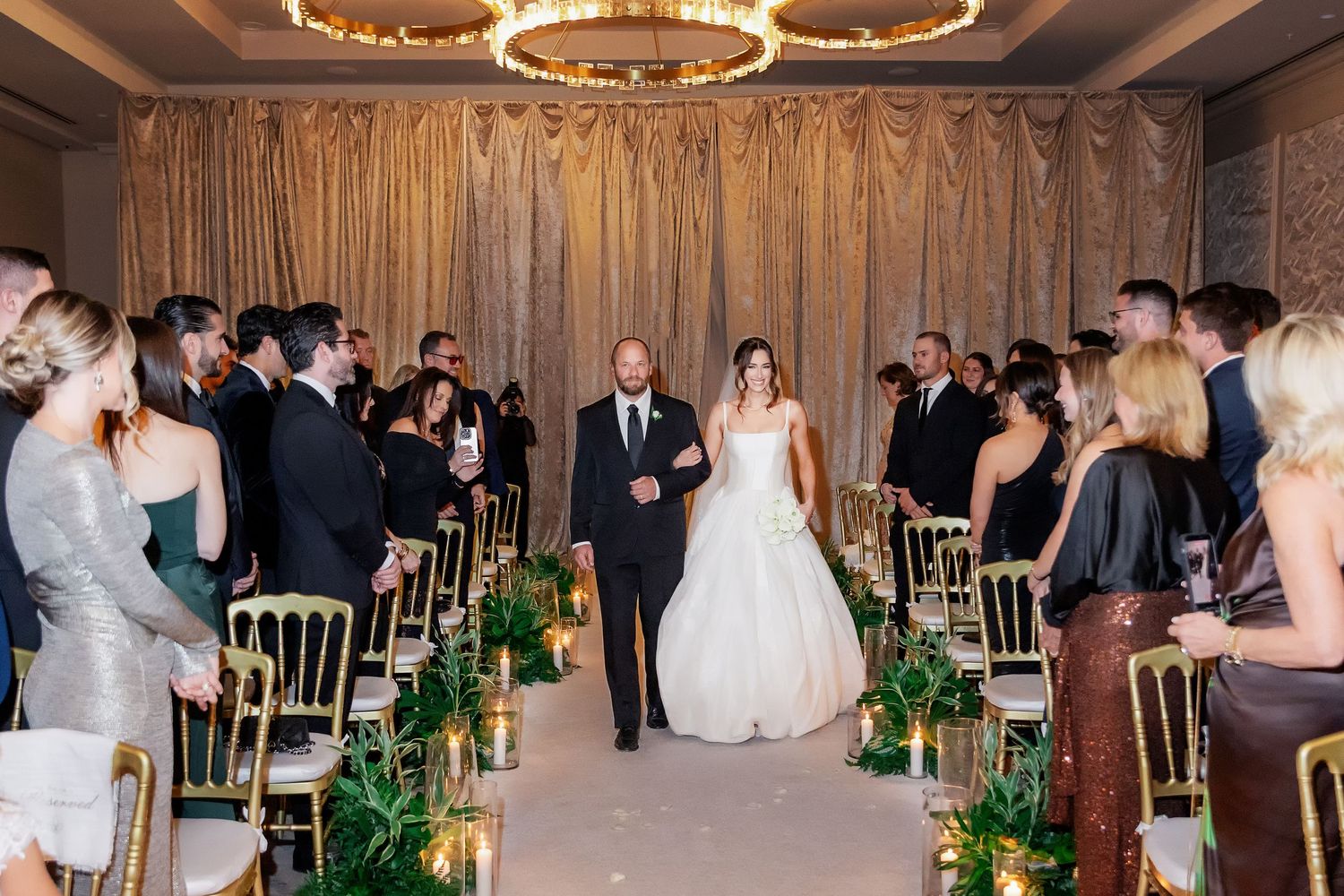 Bride walks down the aisle with escort at elegant indoor wedding ceremony with gold chairs and lush greenery.