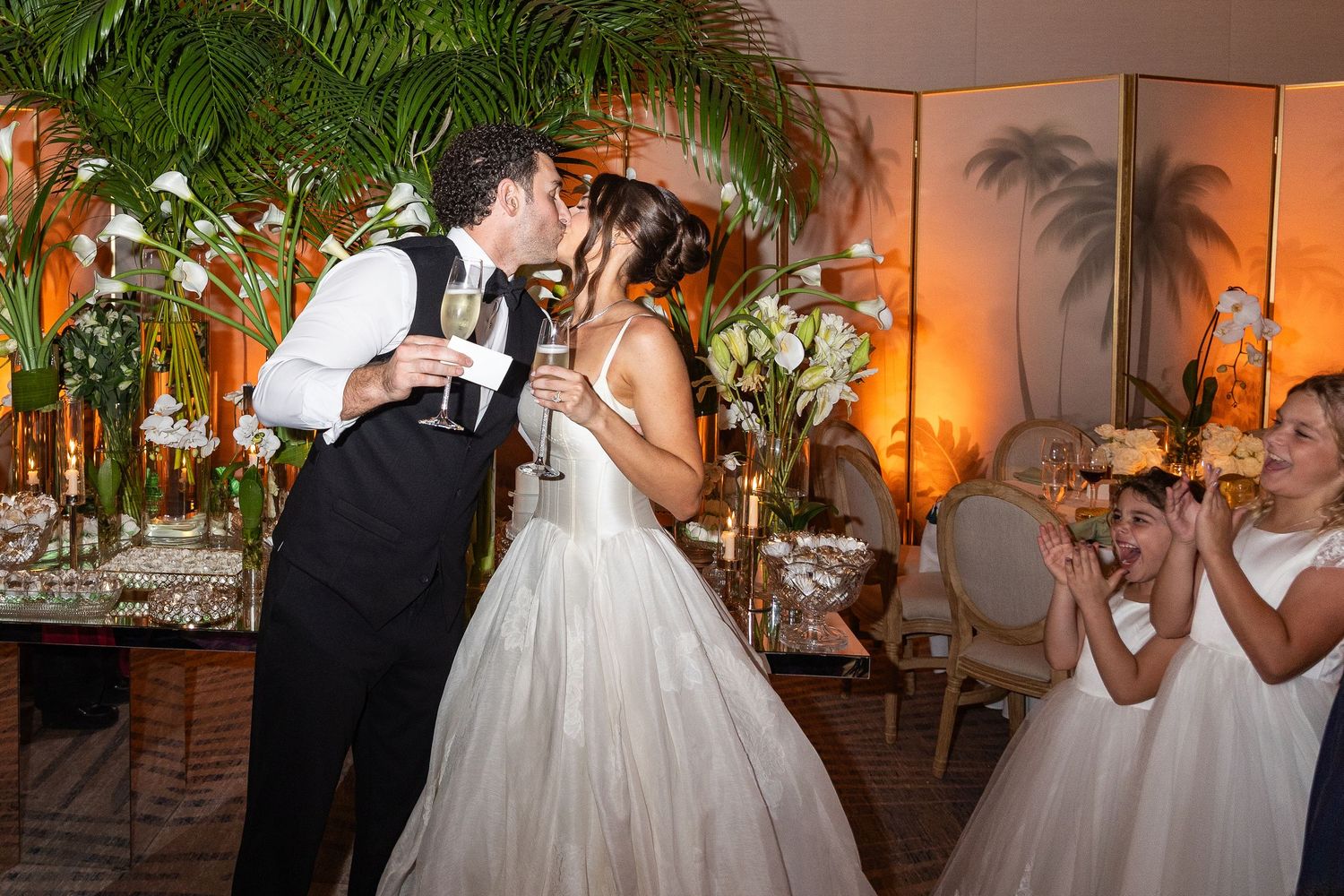 Bride and groom share a kiss while holding champagne glasses at elegant wedding reception with floral decor.