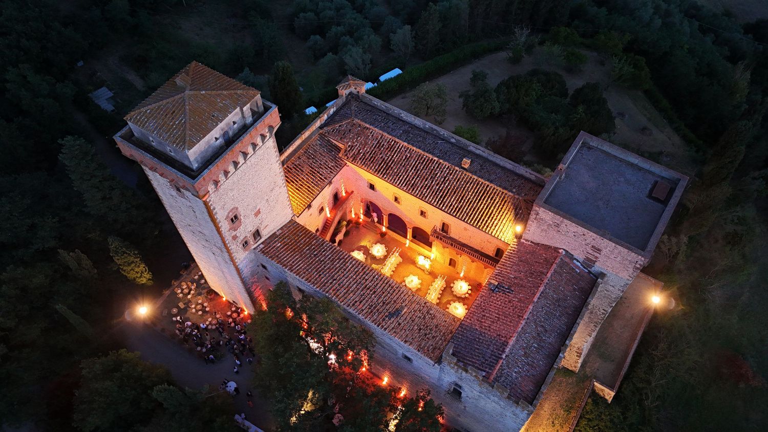Aerial night view of illuminated historic castle with warm glowing courtyard surrounded by lush trees at dusk.