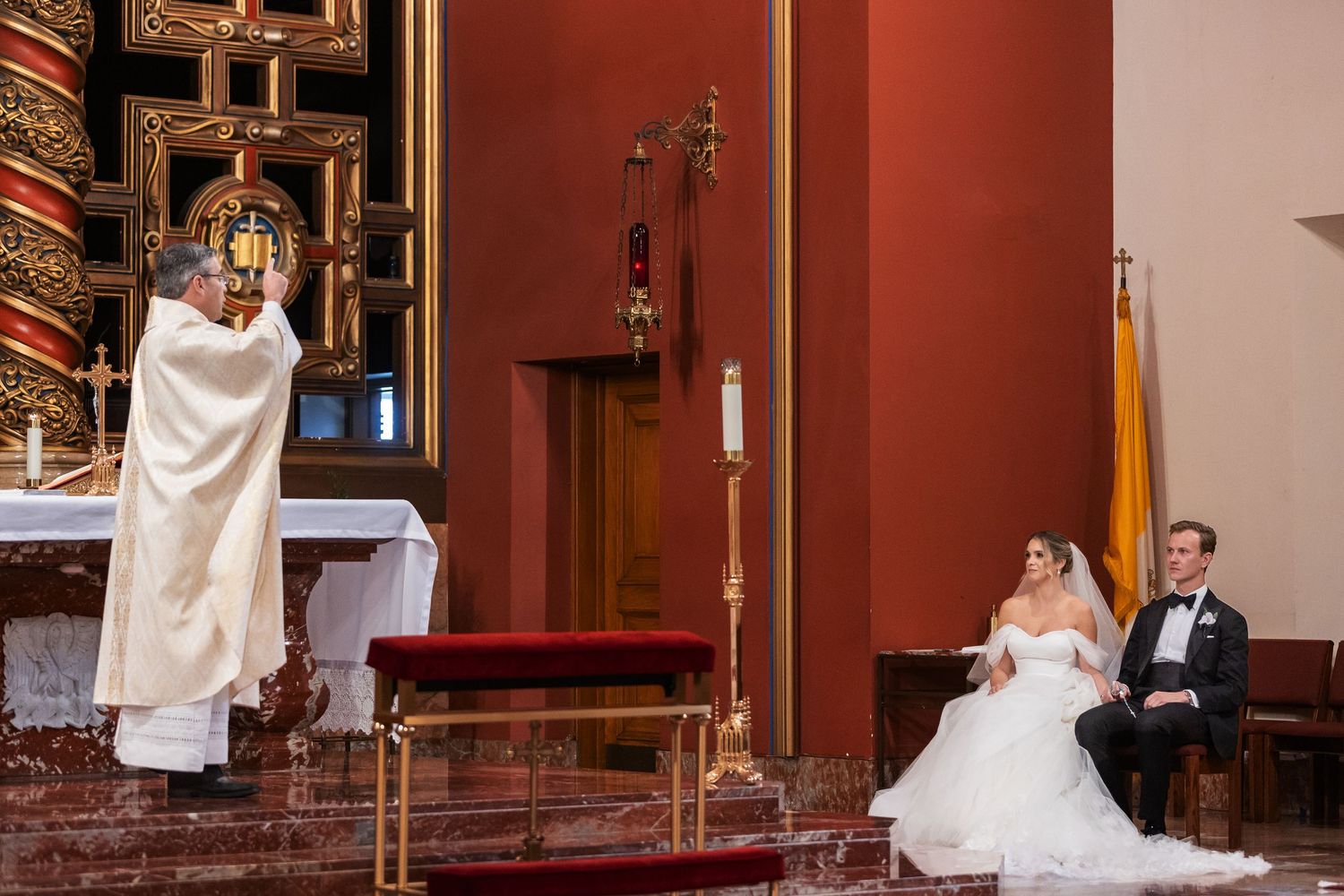 Bride and groom seated at church altar during wedding ceremony while priest performs sacred rites.