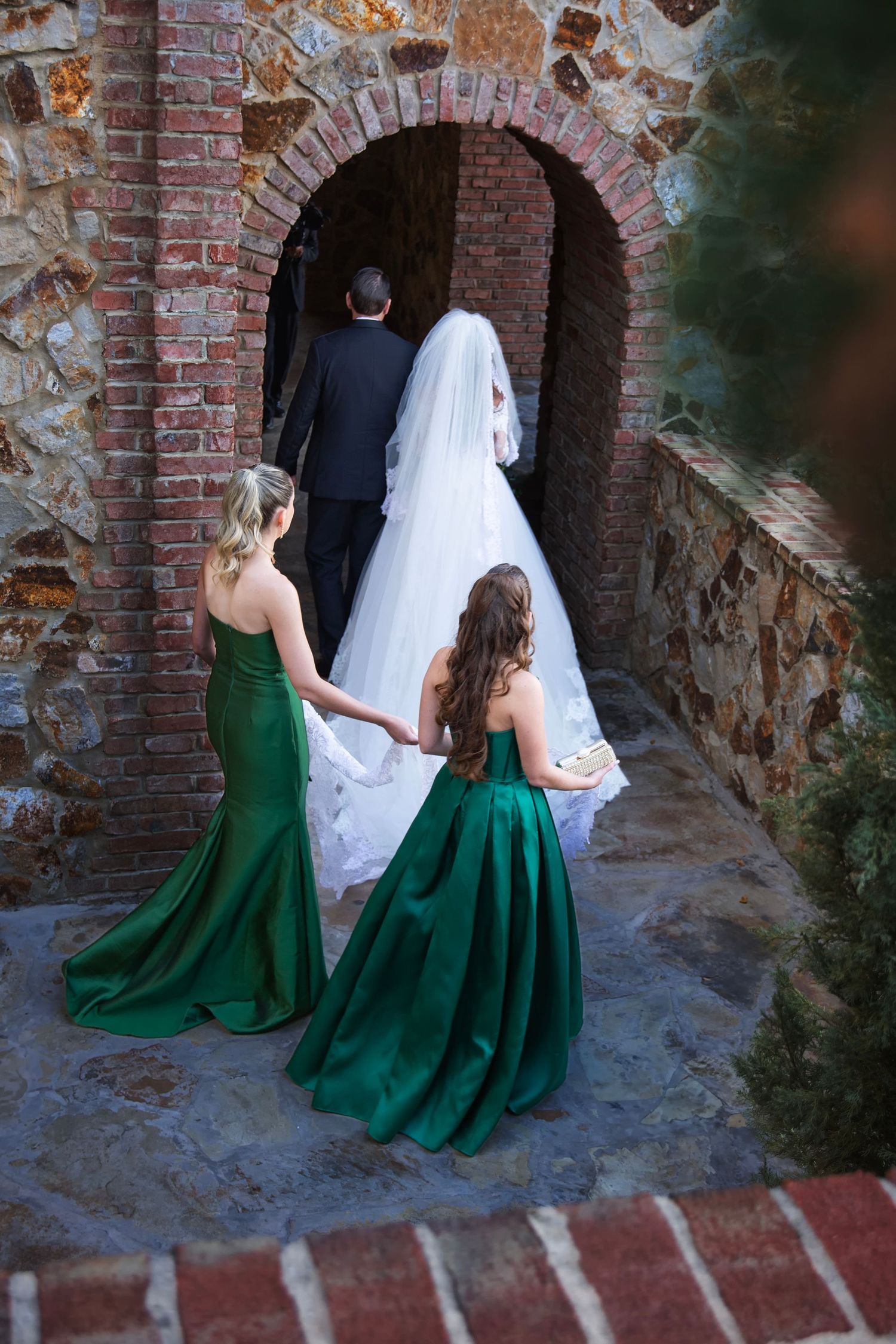 Two bridesmaids in emerald green gowns follow a bride and groom through a rustic brick archway.