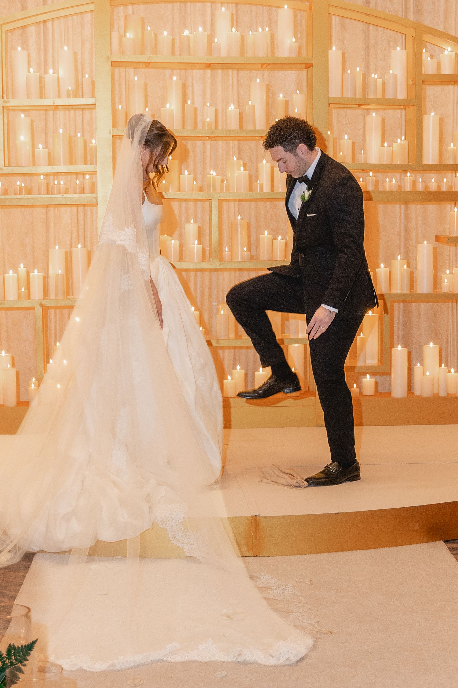 Groom in black tuxedo stomping glass while bride in white ballgown stands before candlelit altar at Jewish wedding ceremony.