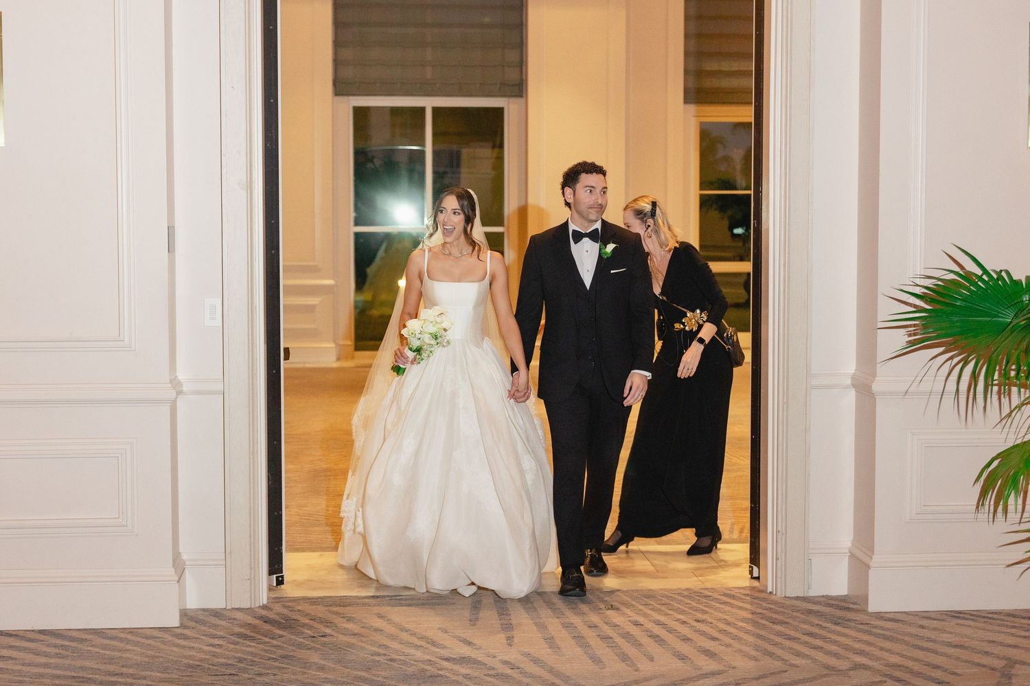 Bride and groom enter wedding venue in elegant white gown and tuxedo, accompanied by a guest in floral dress.