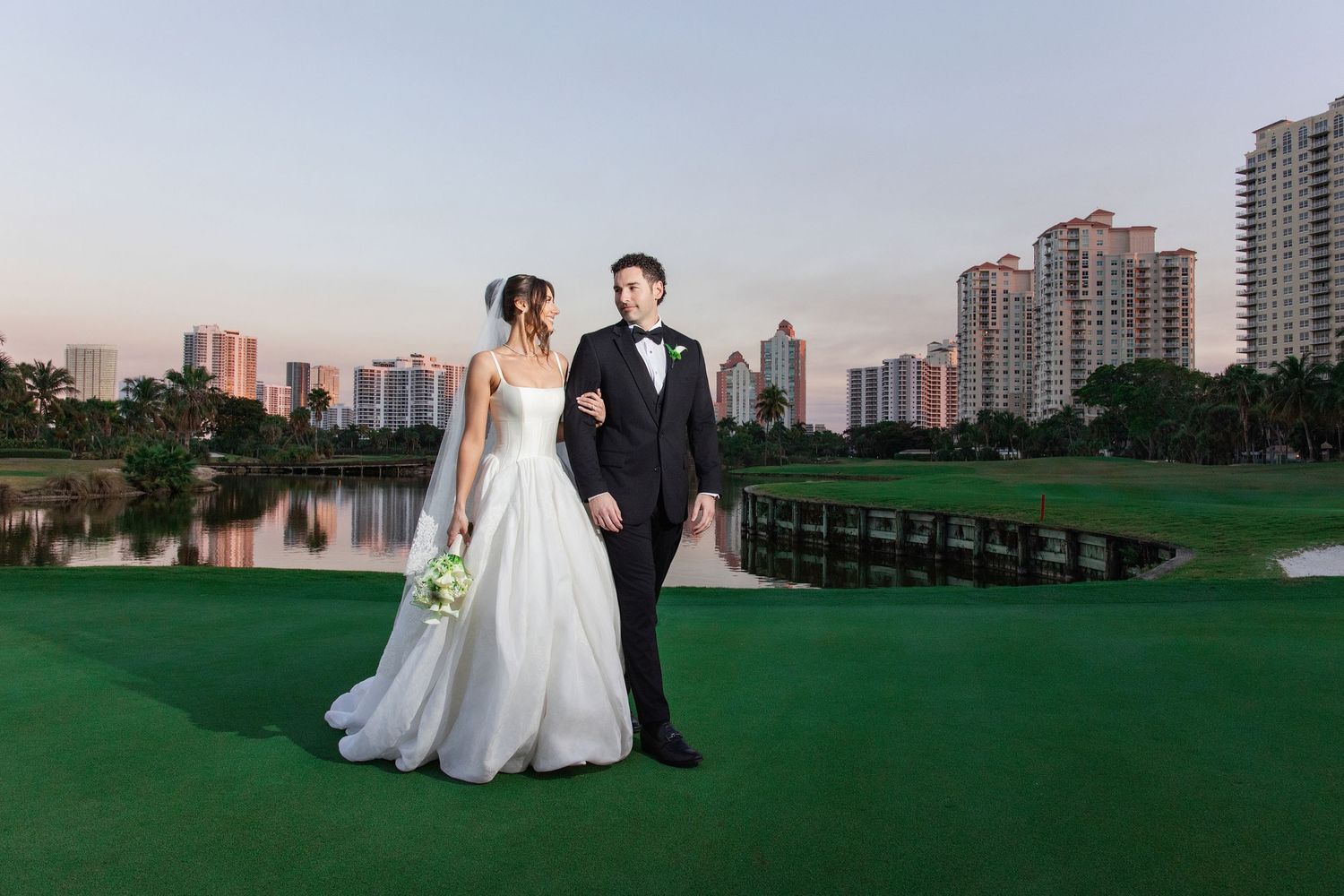 A wedding couple poses in front of a scenic golf course with high-rise buildings in the background at sunset.