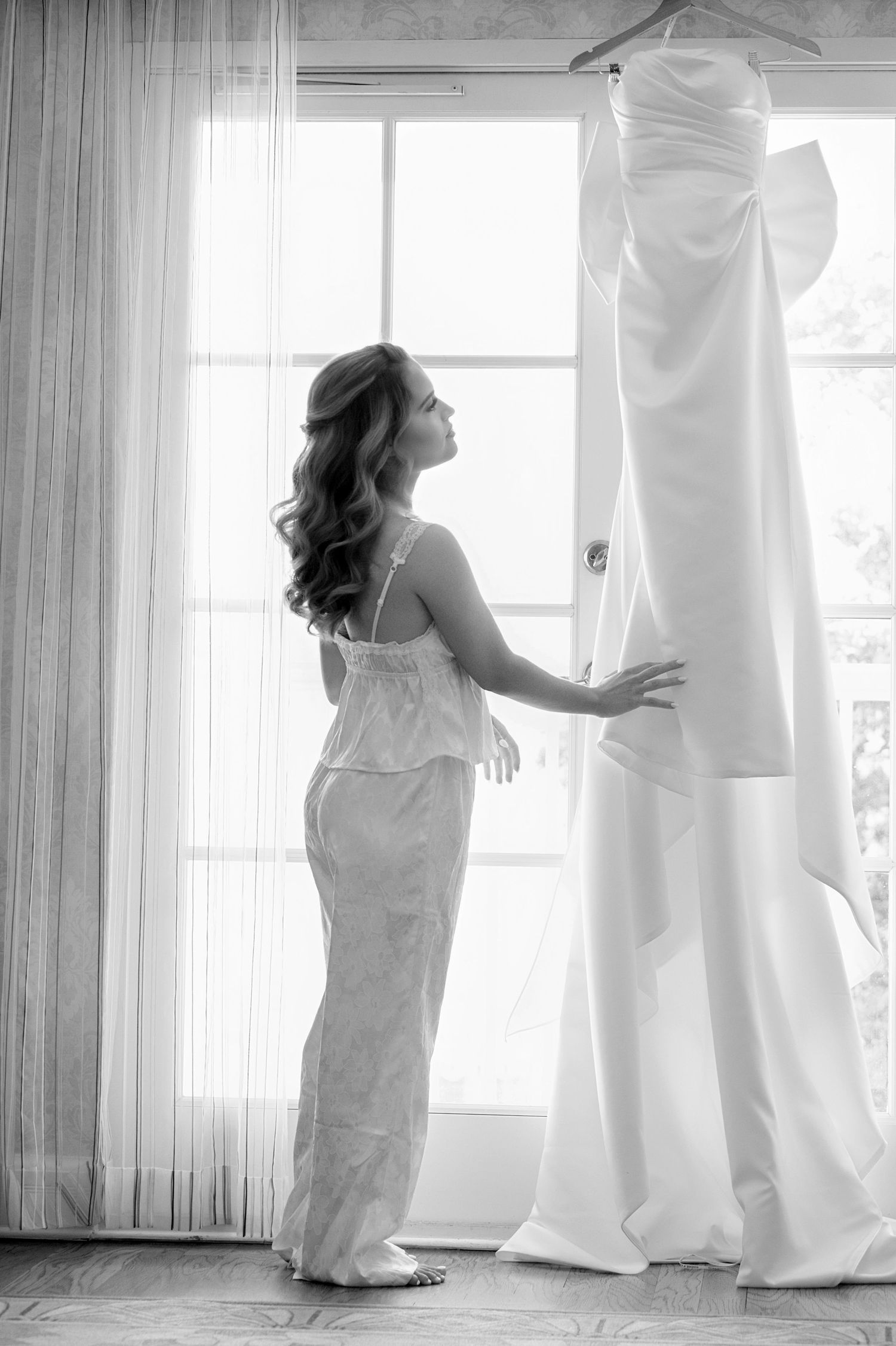 Black and white photo of someone admiring a wedding dress hanging by a window.