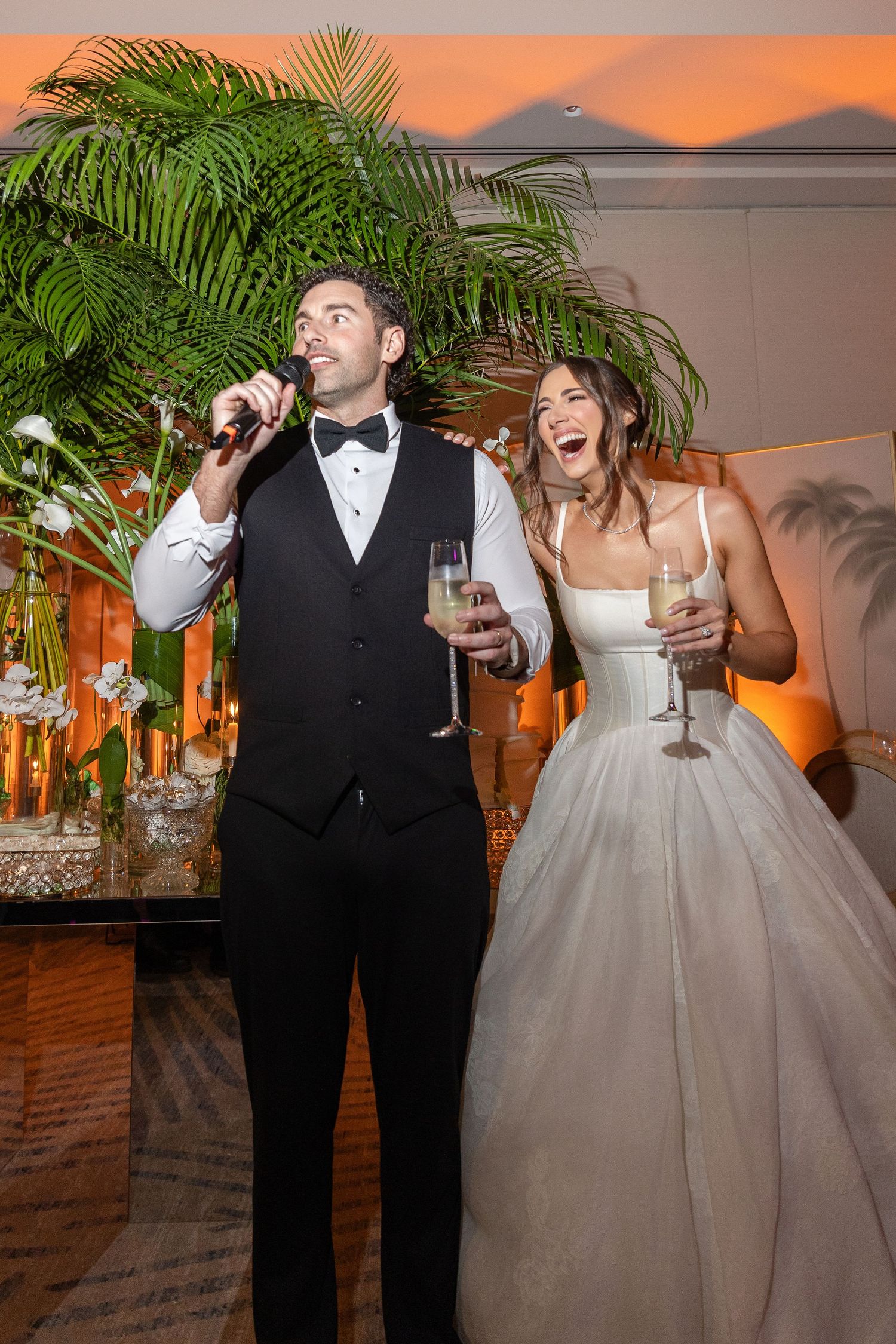 Joyful bride and groom holding champagne glasses, groom speaking into microphone at elegant wedding reception with tropical decor.