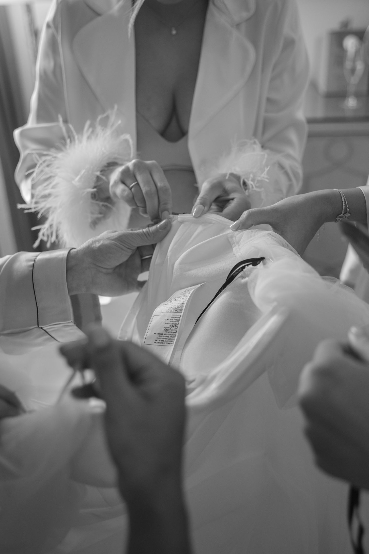 Black and white photo of bridesmaids helping a bride into her white wedding dress with feather details.