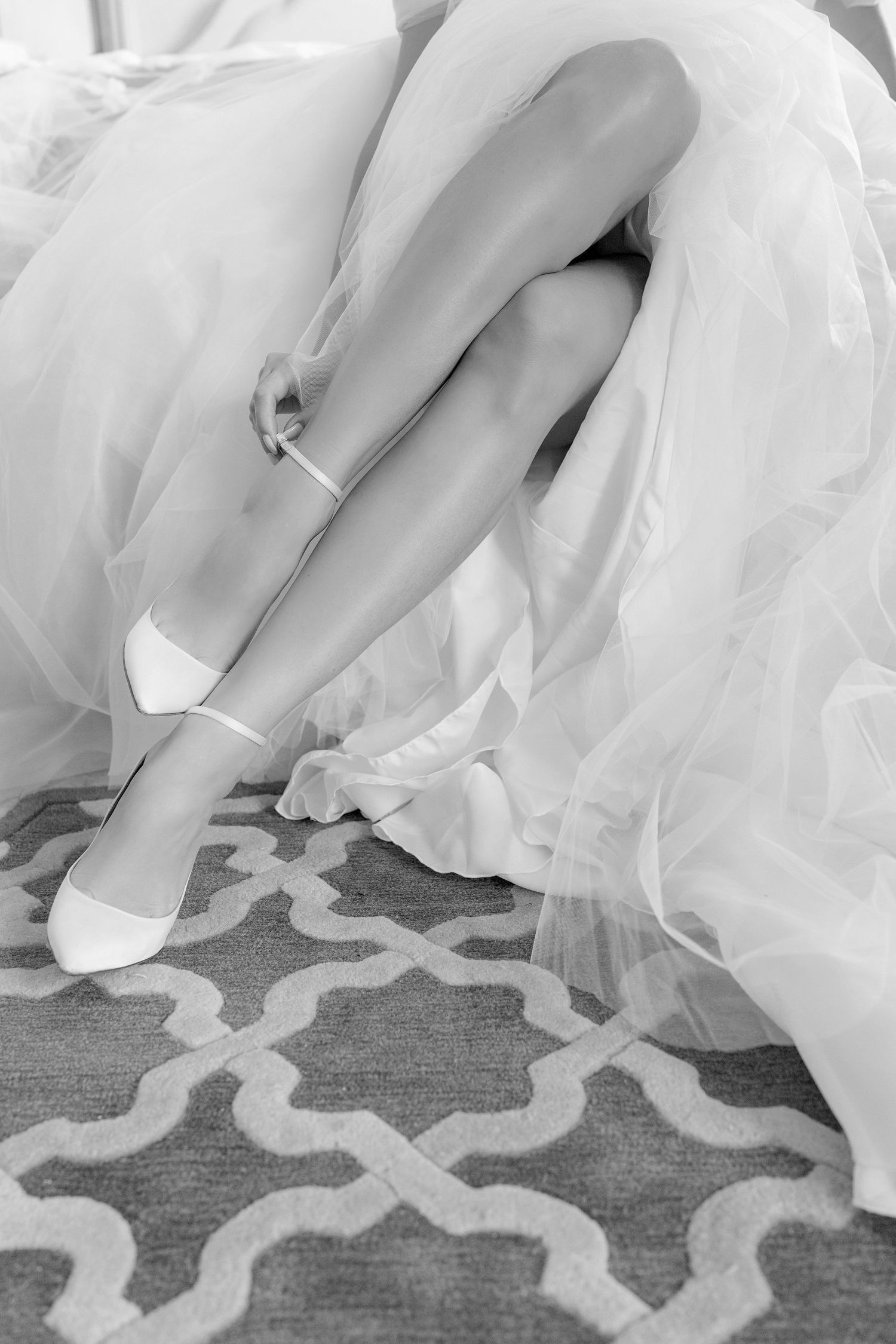 Black and white photo of a bride in a white gown adjusting her elegant white heels on a patterned carpet.