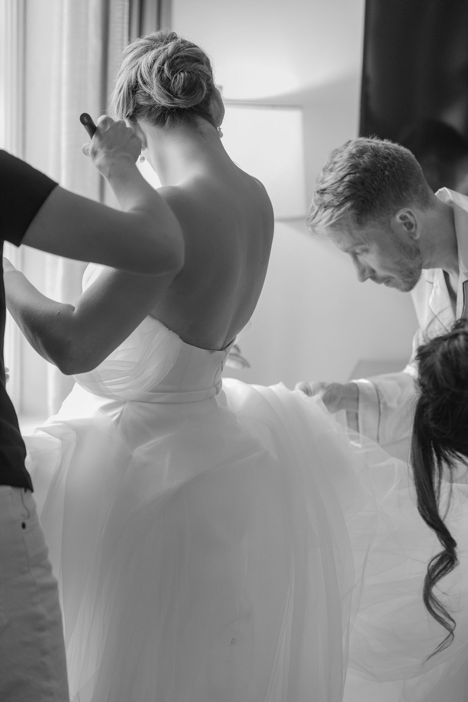 Black and white photo of a bride in a backless wedding gown being helped by attendants before her ceremony.