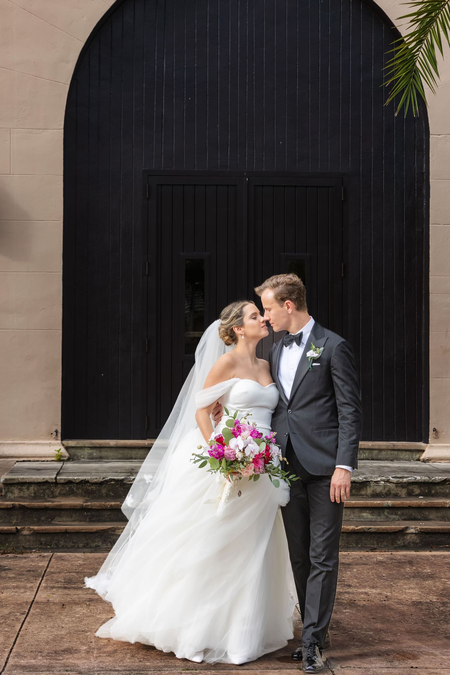 Bride and groom share a kiss in front of a black arched door, bride holding a vibrant pink and white bouquet.