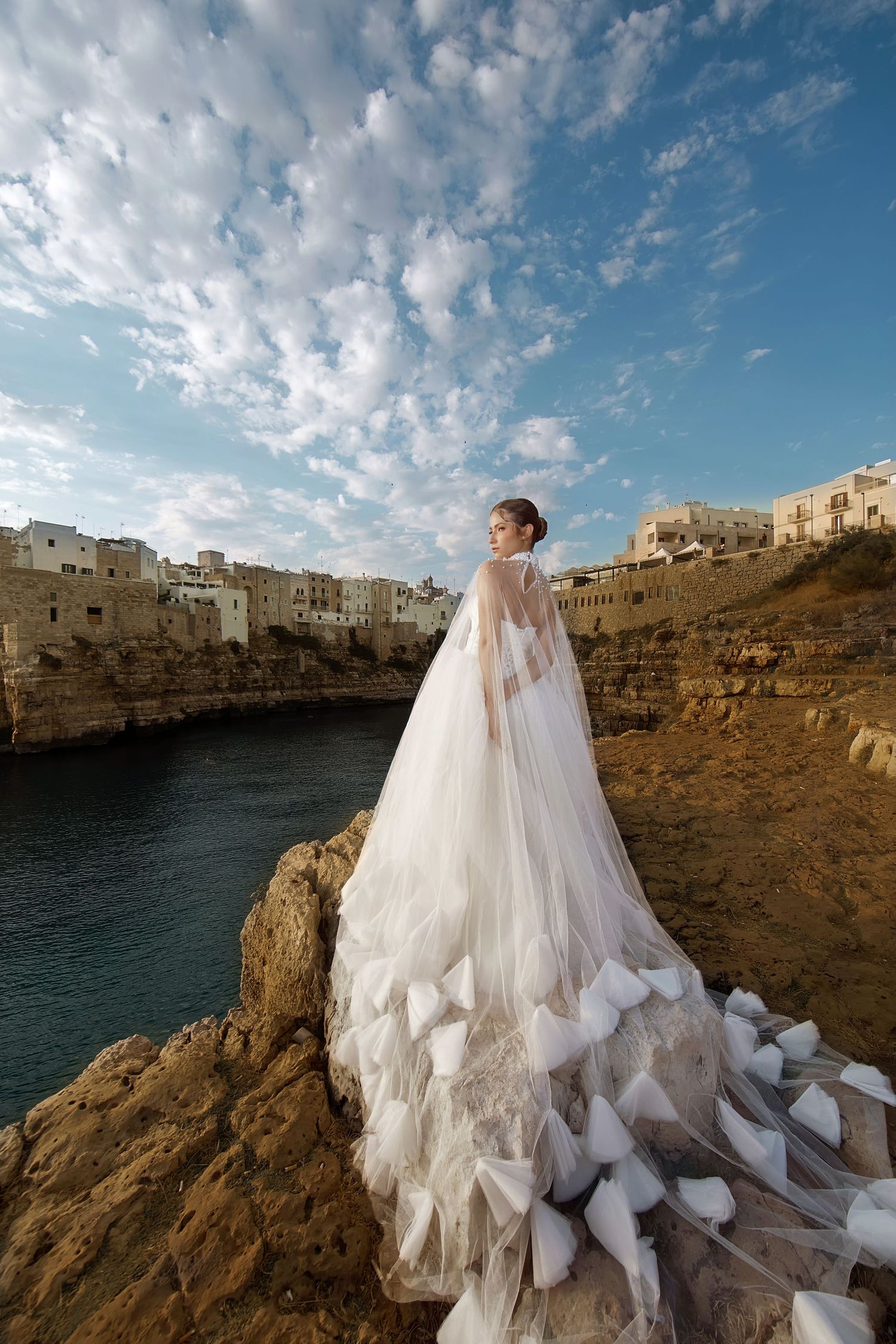 Bride in elegant white gown with flowing tulle cape stands on rocky cliffs overlooking a coastal Italian town under blue skies.
