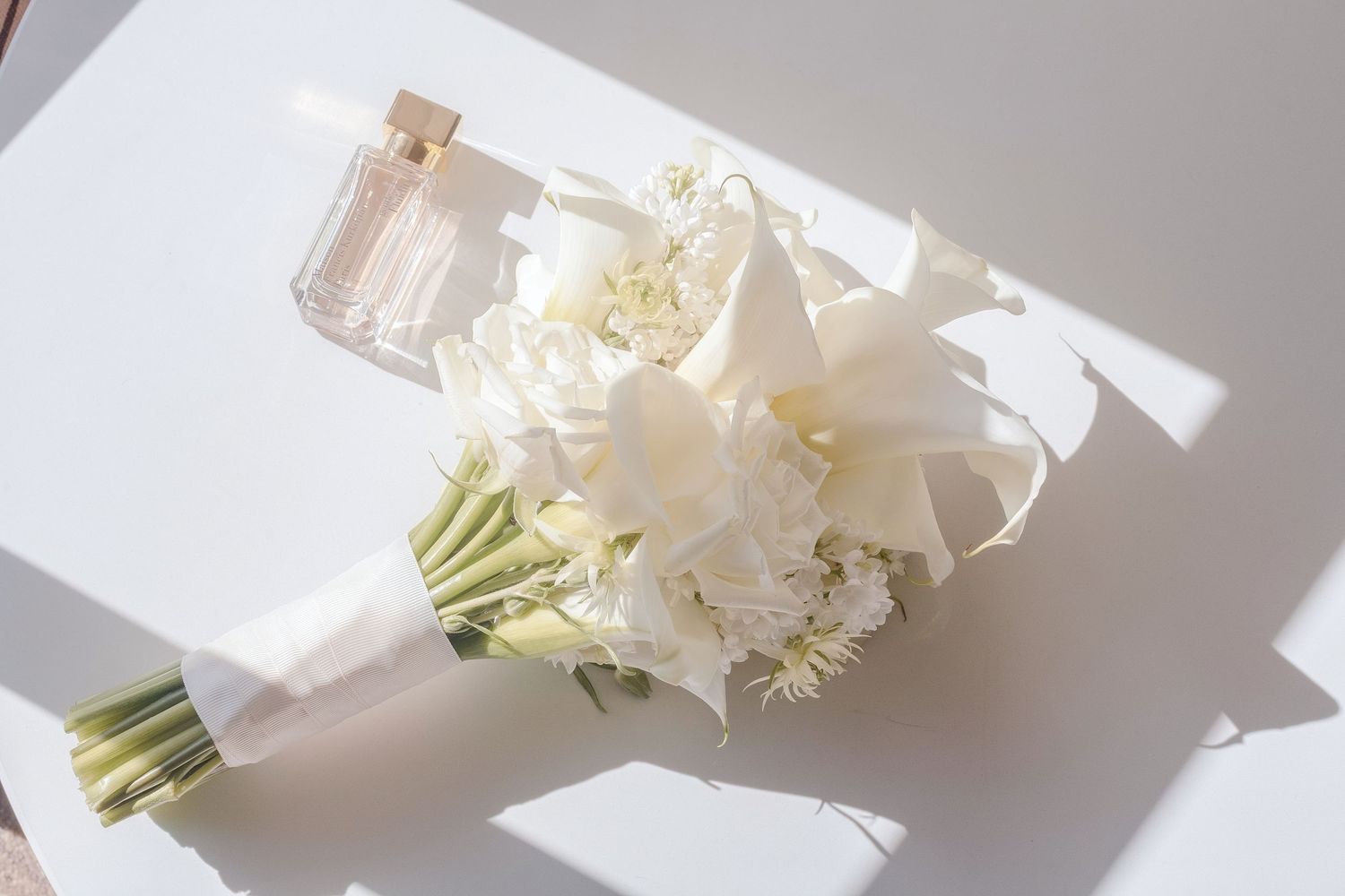 White wedding bouquet with lilies and carnations beside a pink perfume bottle on a white surface.