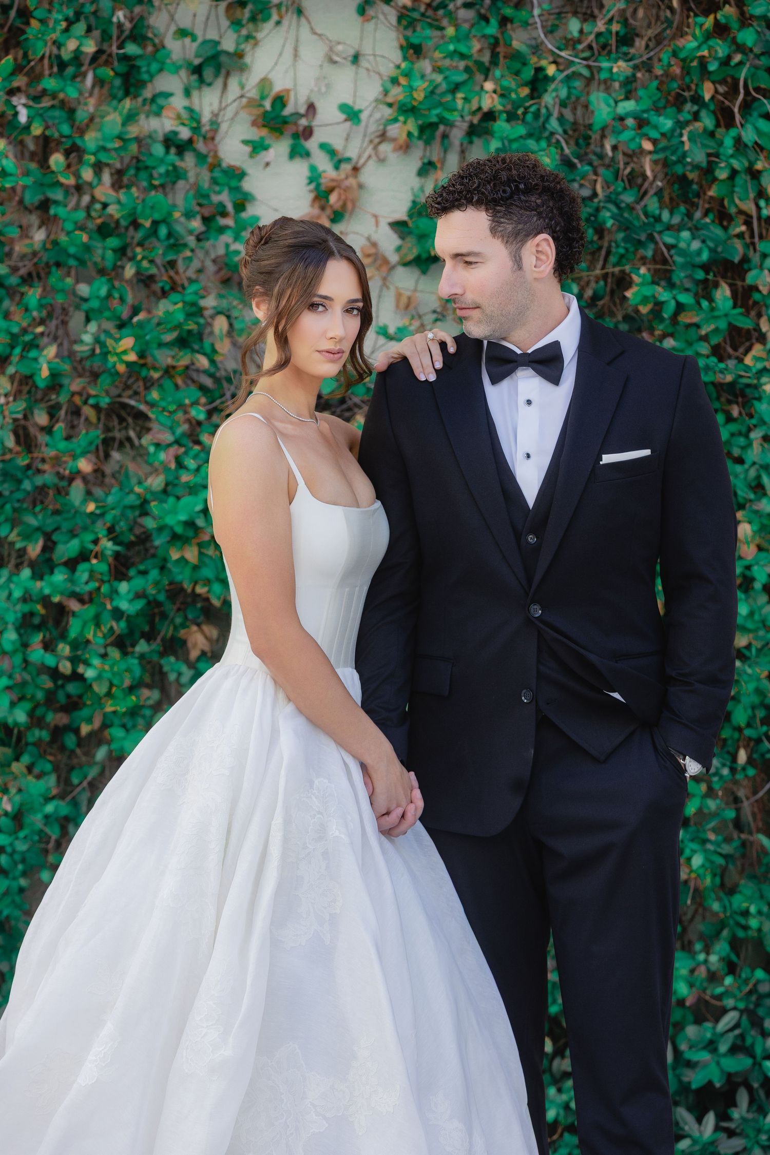 Bride in white ball gown and groom in black tuxedo pose together before a lush green ivy wall backdrop.