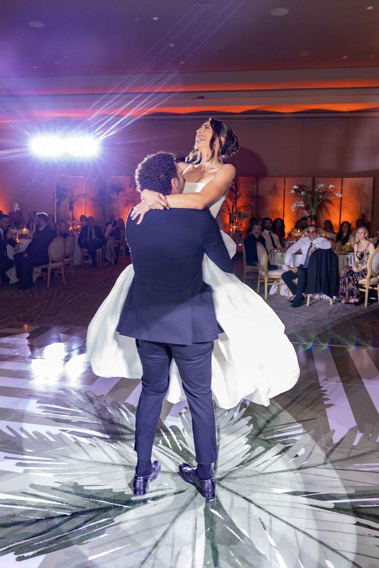 Groom lifts bride during first dance at elegant wedding reception with warm amber lighting and guests watching.