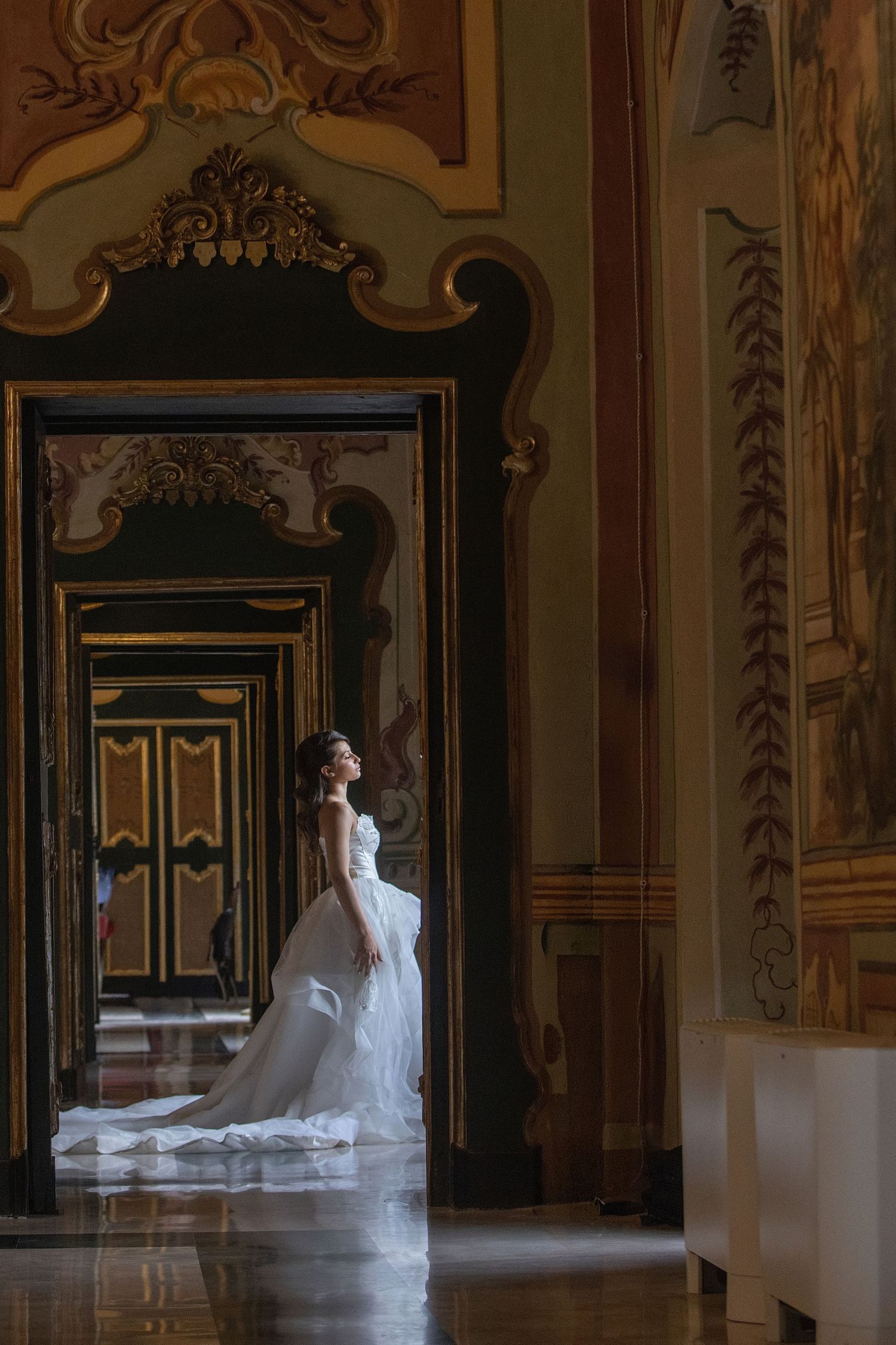 Bride in white gown stands in ornate baroque palace hallway with gilded doors and frescoed walls.