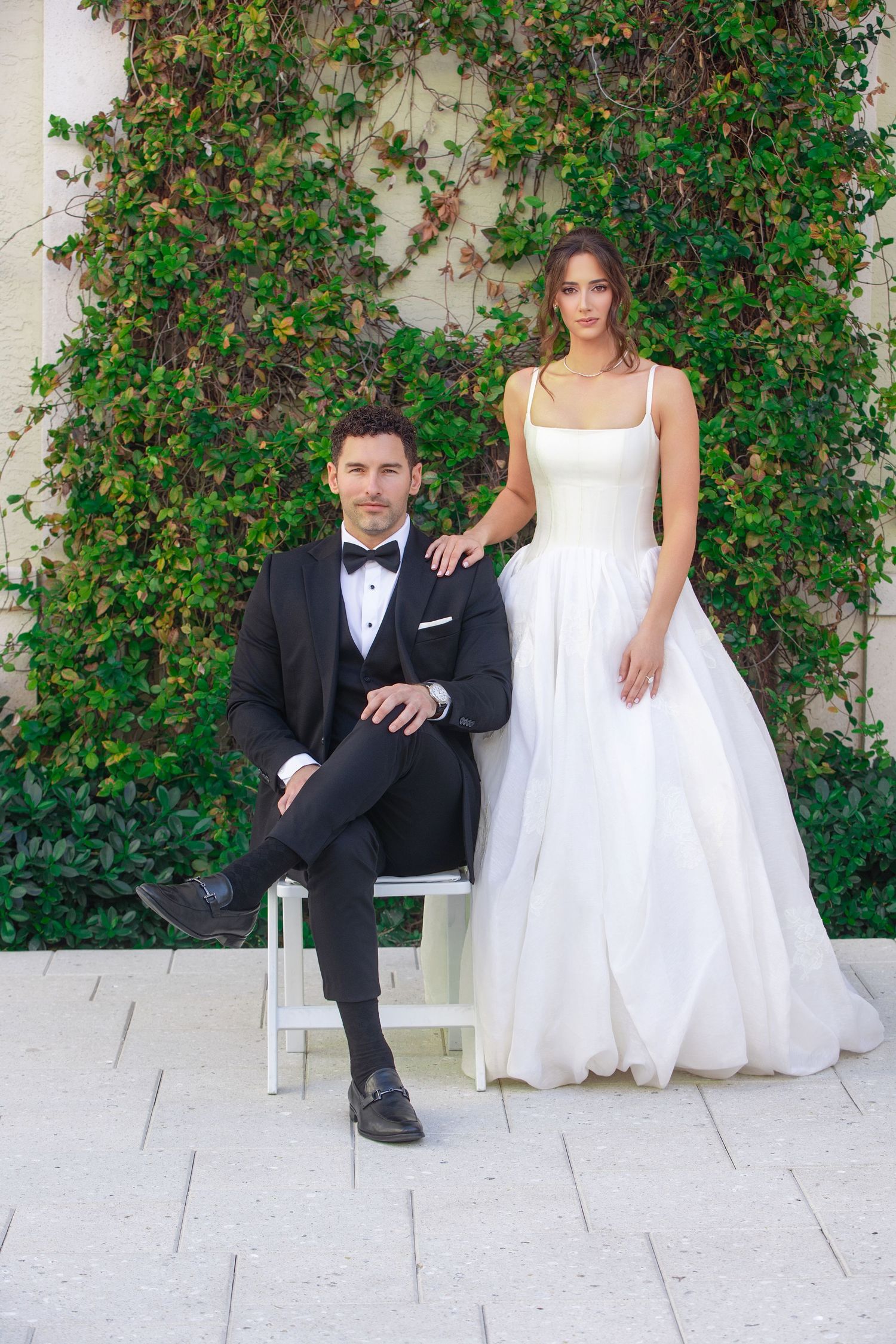 A wedding couple poses casually on white stone steps against a lush green vine-covered wall.