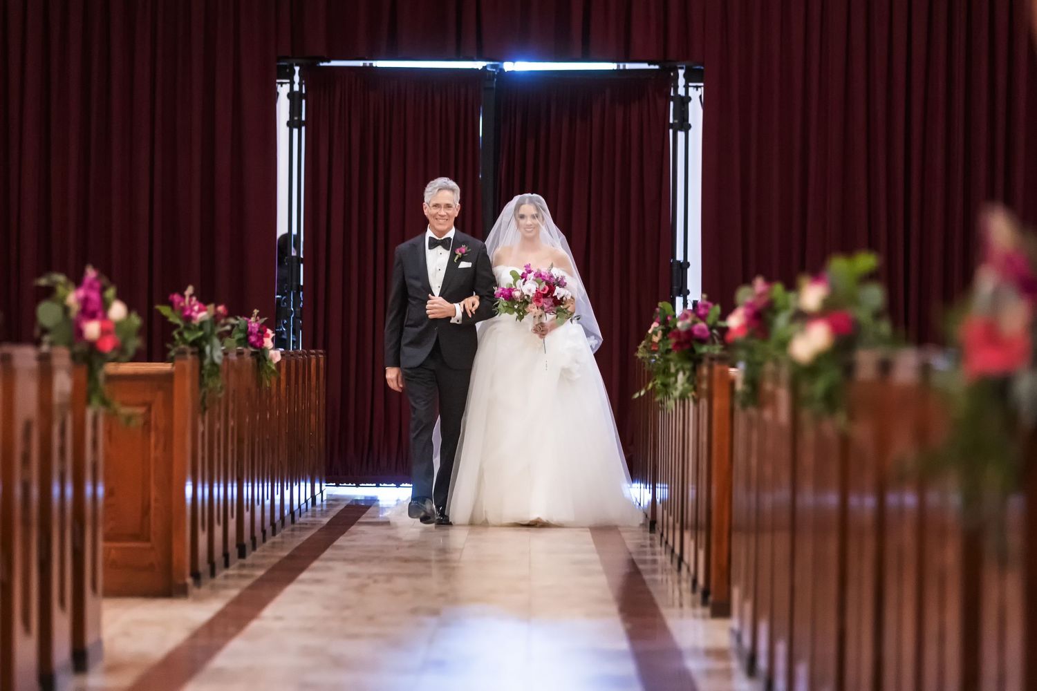 Bride and father walking down church aisle lined with pink floral arrangements during wedding ceremony.