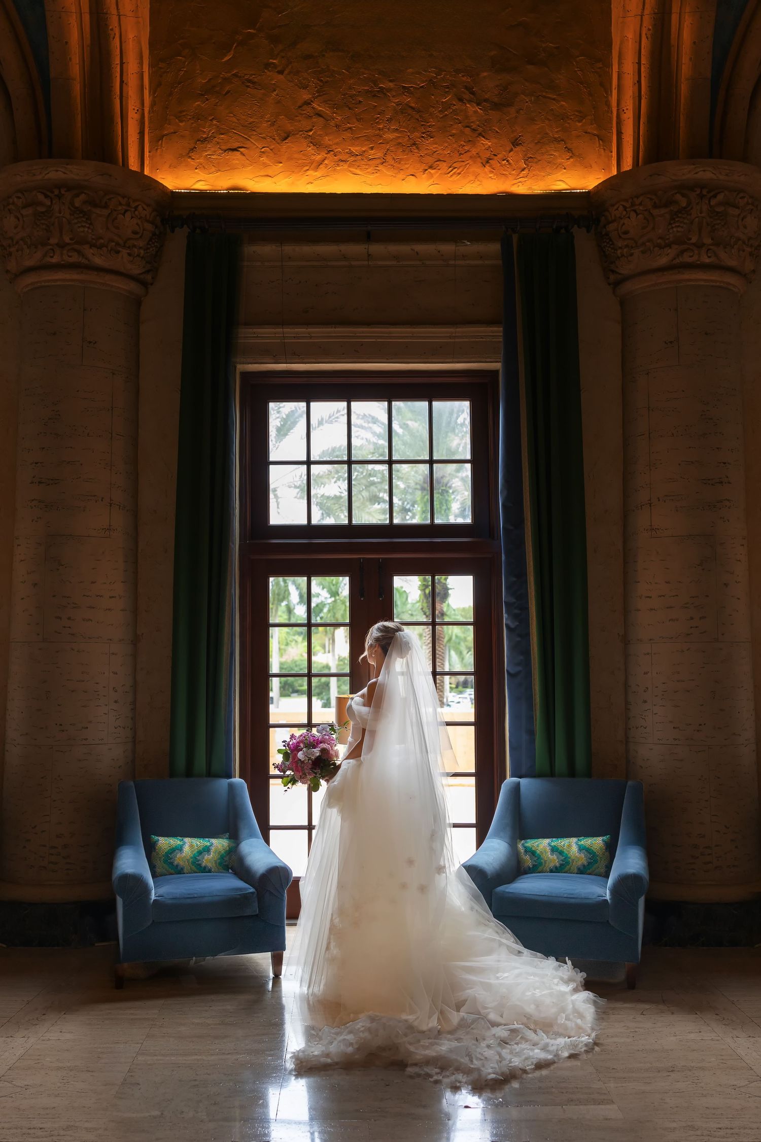 Bride in white gown and veil stands before tall window between blue chairs in elegant ornate venue.