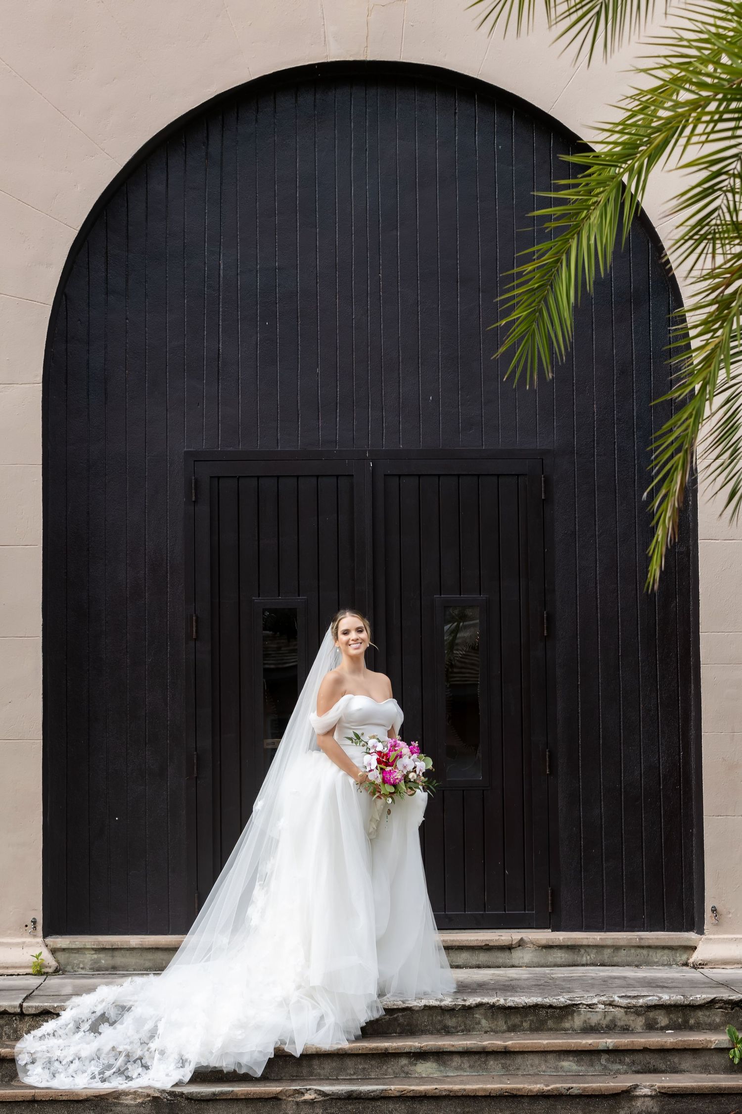 Bride in white ball gown and veil holds pink bouquet before grand black arched doors with palm fronds.
