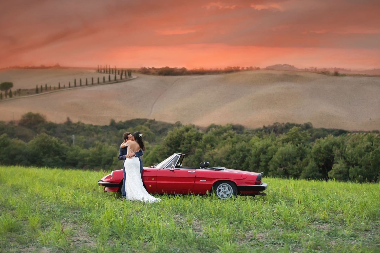 Romantic sunset wedding photo with vintage red car against rolling Tuscan hills.