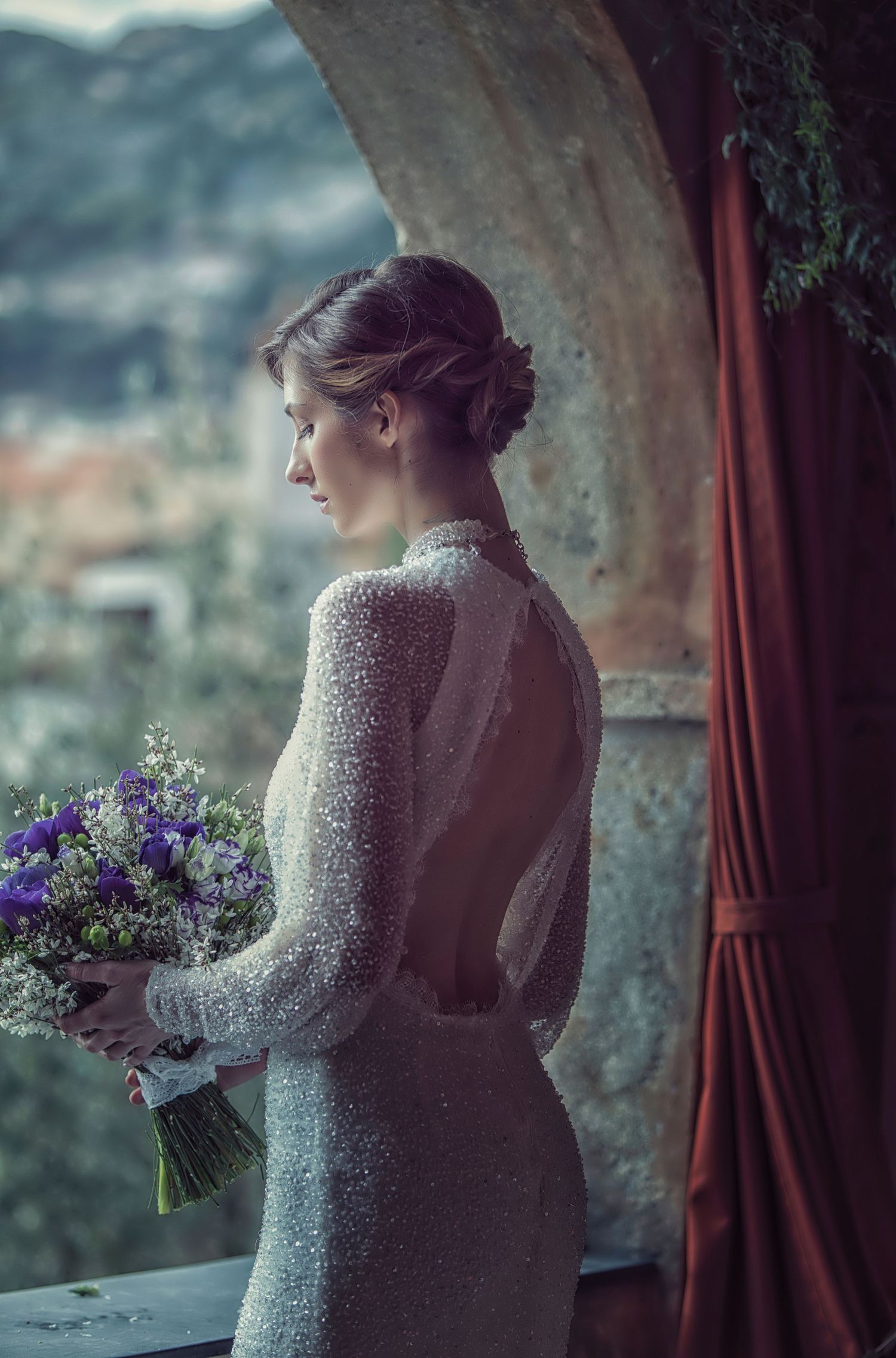Elegant lace wedding dress silhouette against a window with purple flowers.