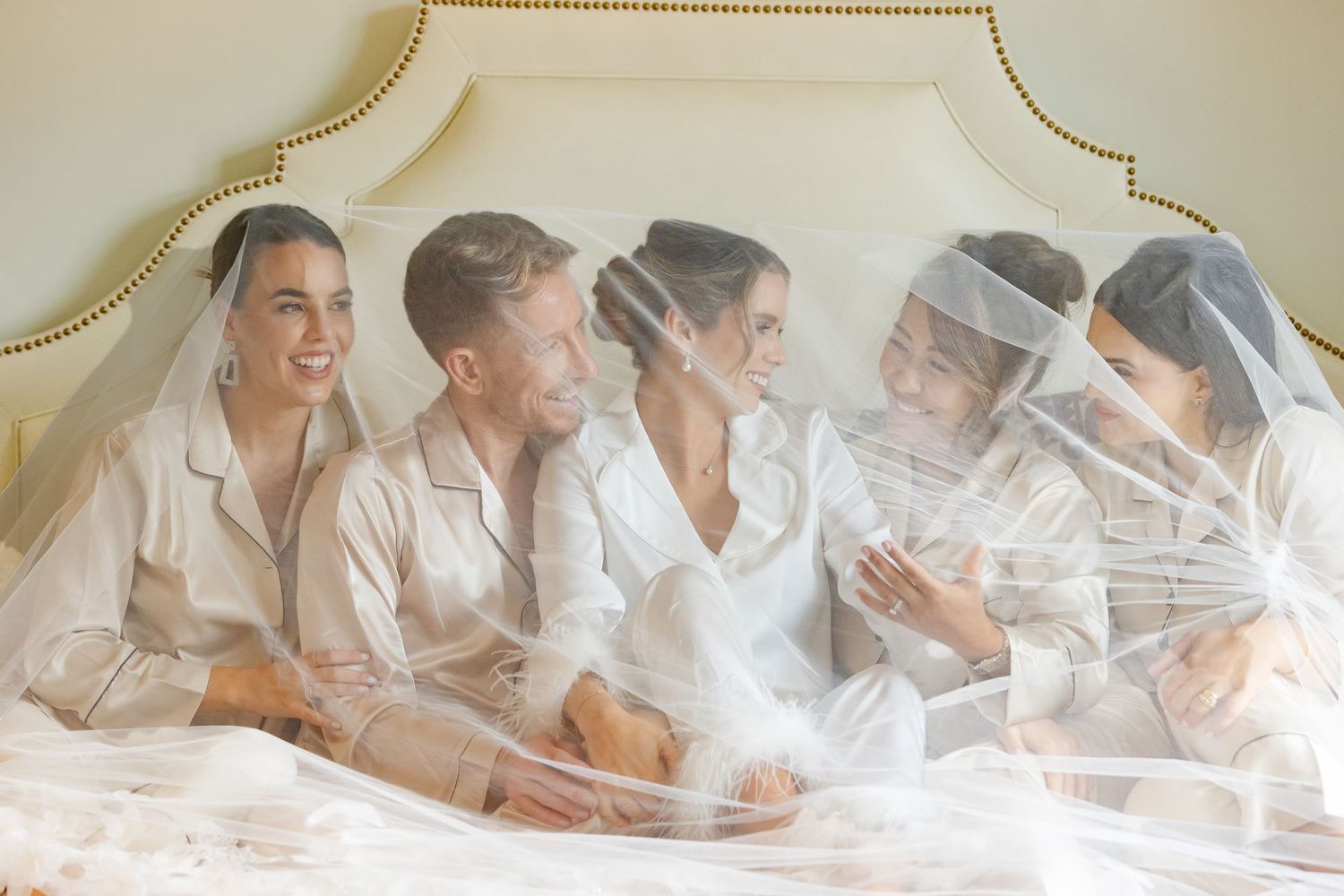 Five bridesmaids in satin pajamas sit together on a bed under a flowing wedding veil, laughing and celebrating.