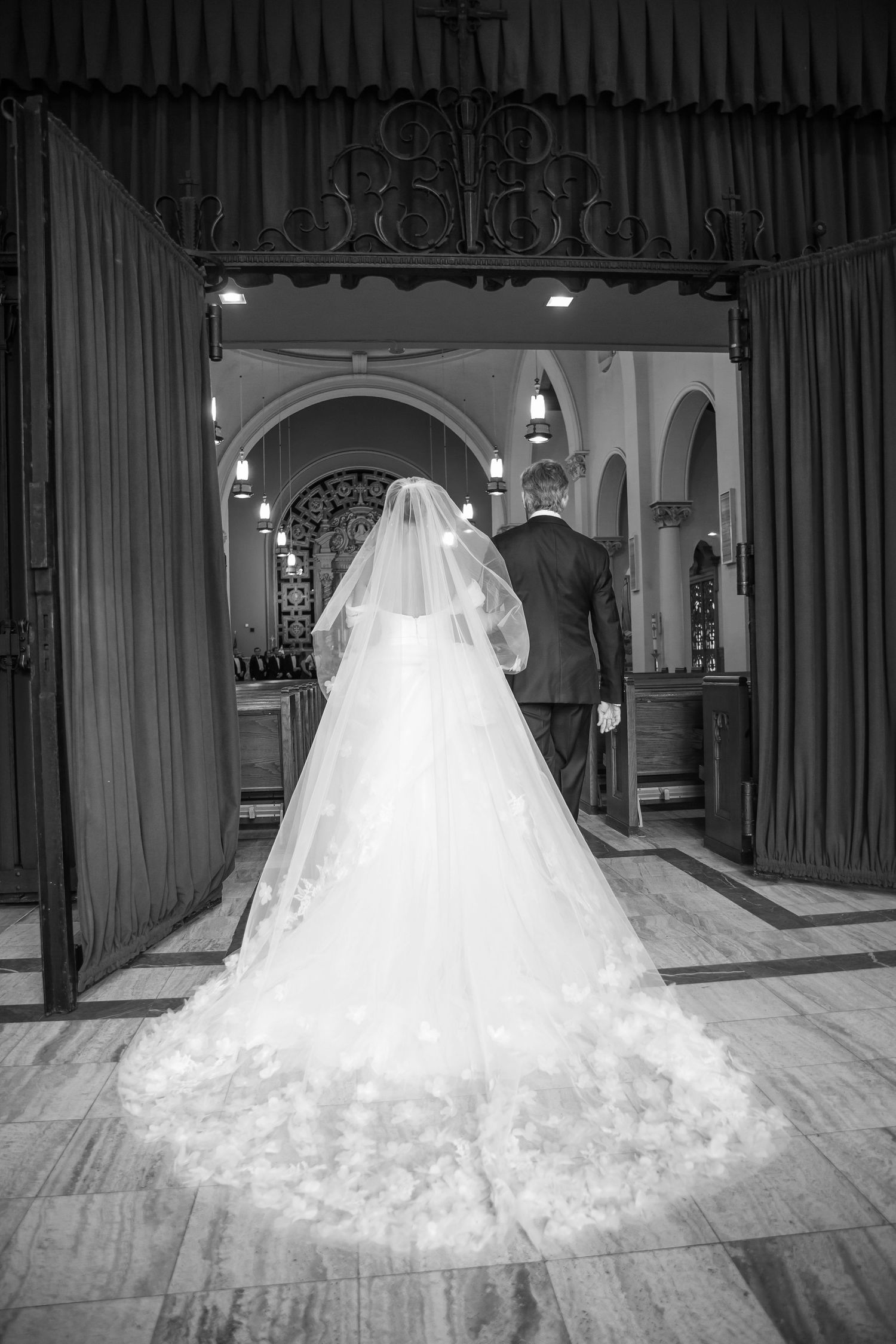 Black and white photo of a bride in a long veil gown entering a church with groom.