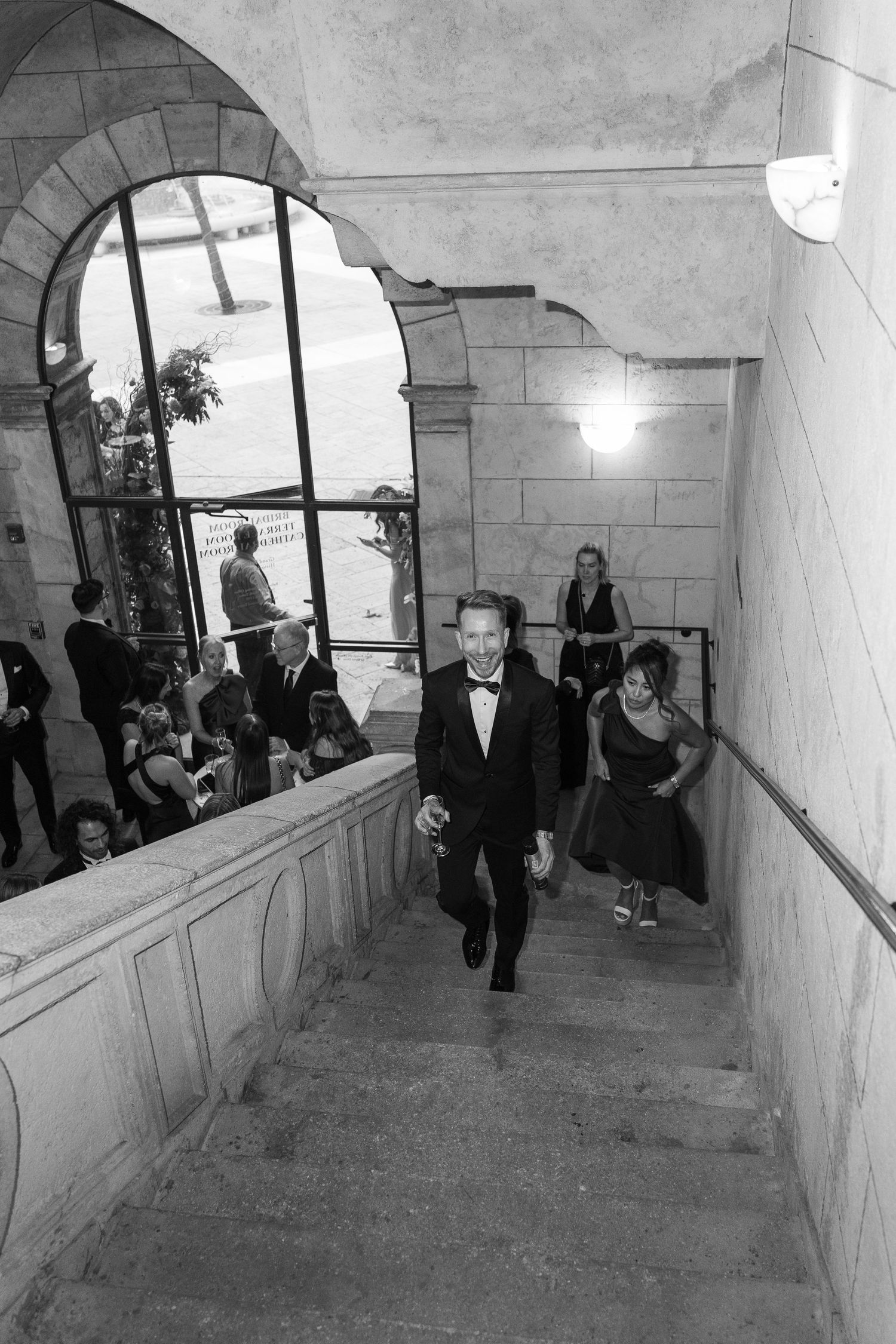 Man in tuxedo smiling while ascending stone stairs at formal event, black and white photography.