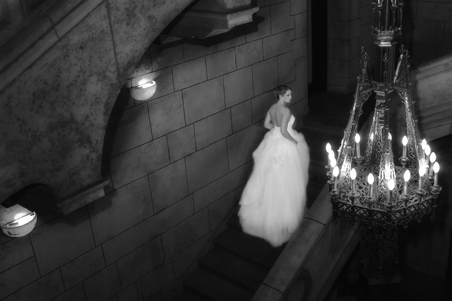 Black and white photo of a bride in a white gown ascending grand stone stairs near an elegant chandelier.