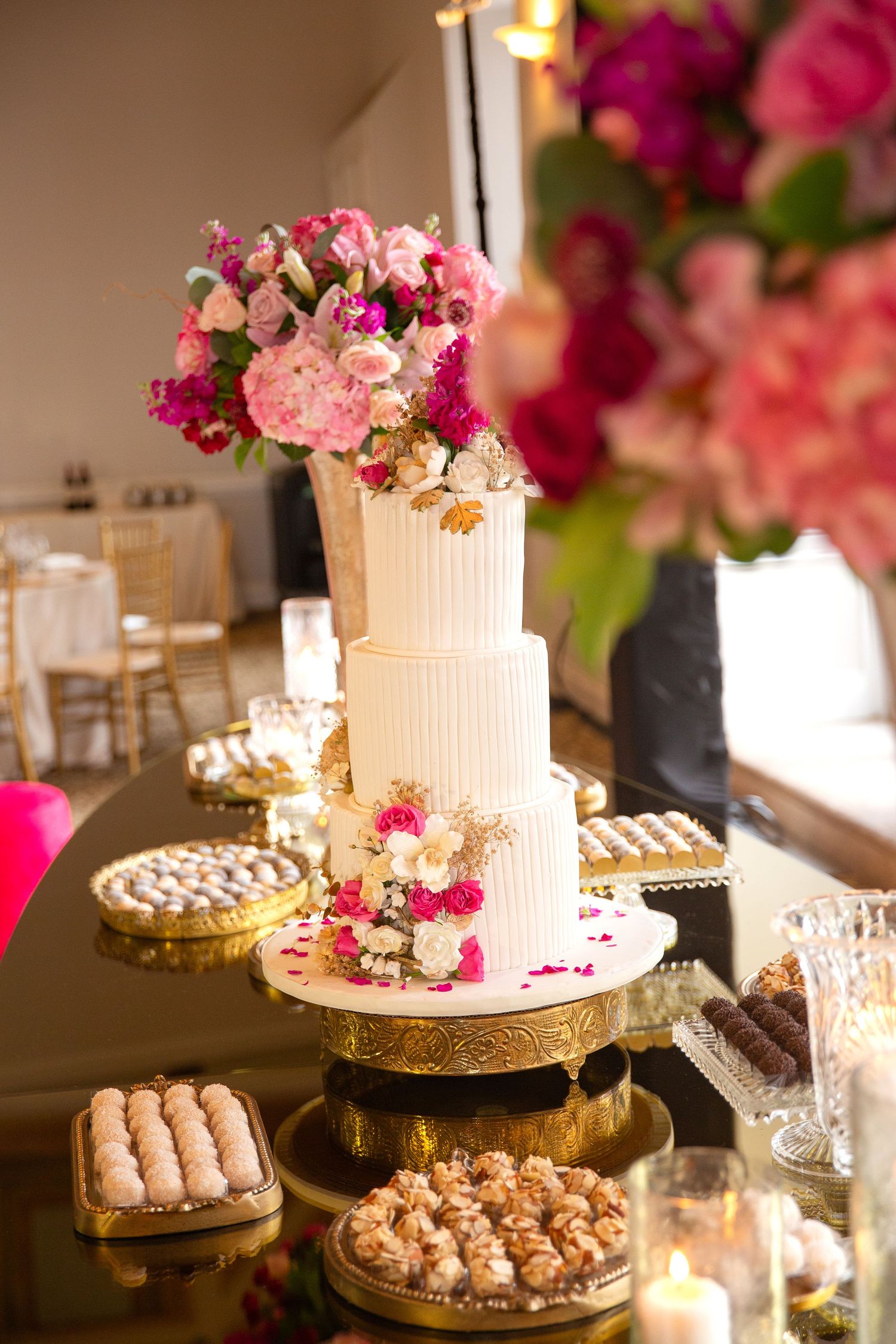 Elegant three-tier white wedding cake adorned with pink flowers on a gold stand, surrounded by desserts and floral arrangements.
