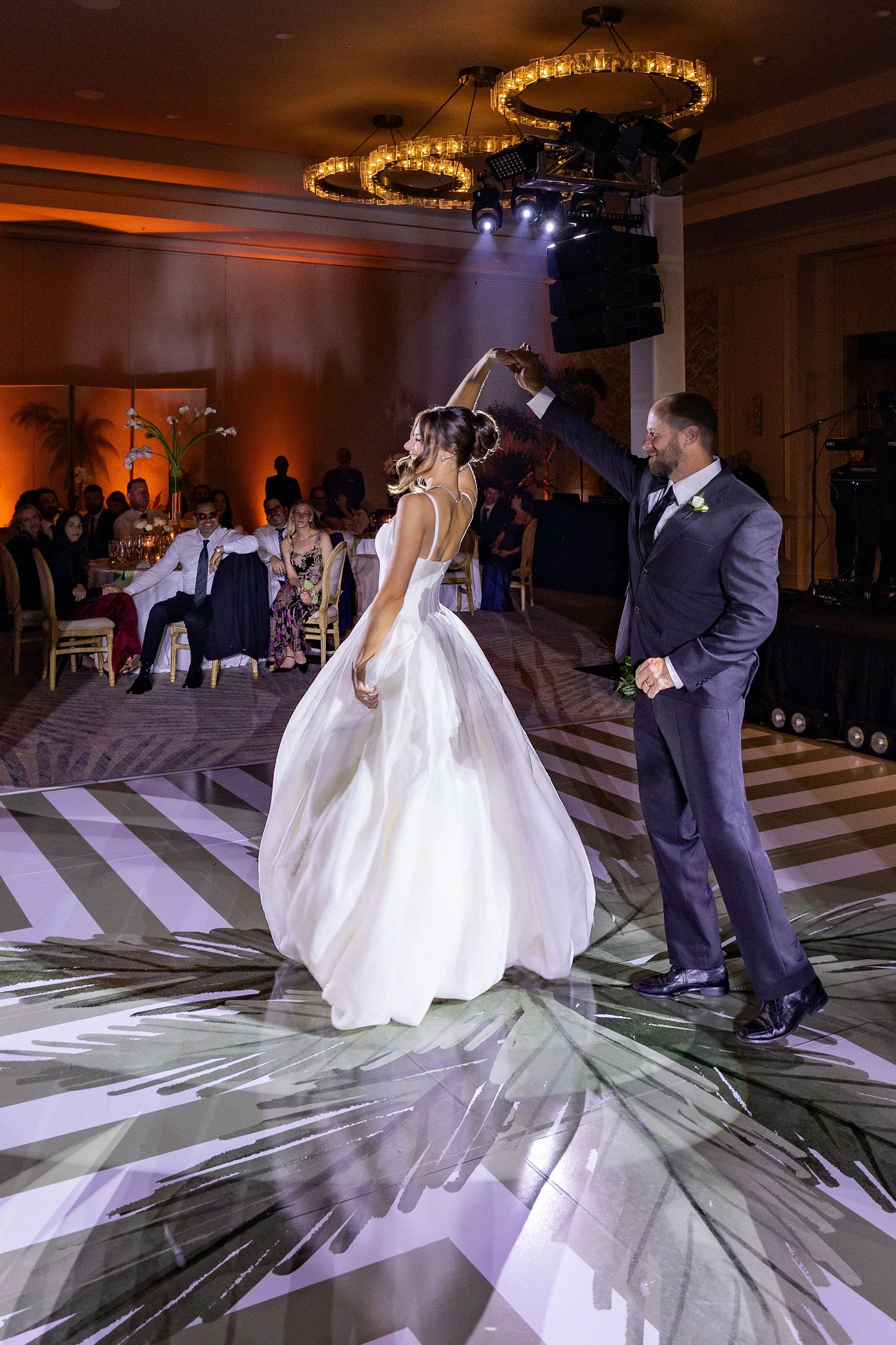 Bride and groom share their first dance on a patterned dance floor in an elegantly lit wedding reception ballroom.
