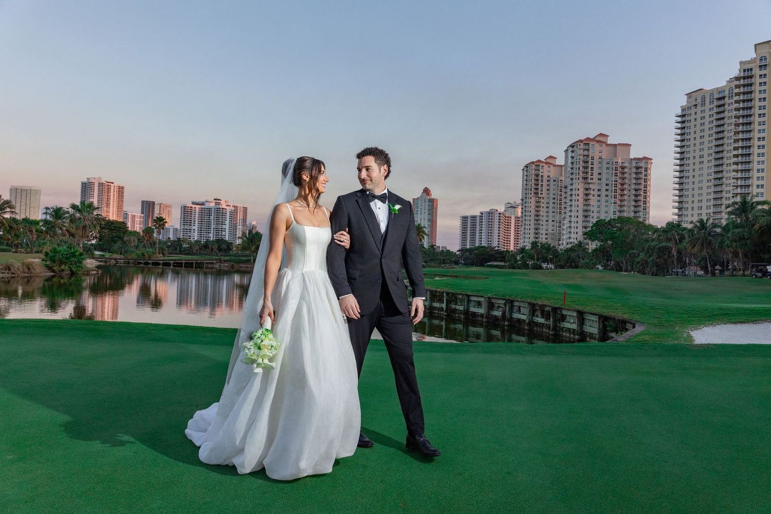 Bride and groom pose on a lush golf course at sunset with a city skyline and lake in the background.
