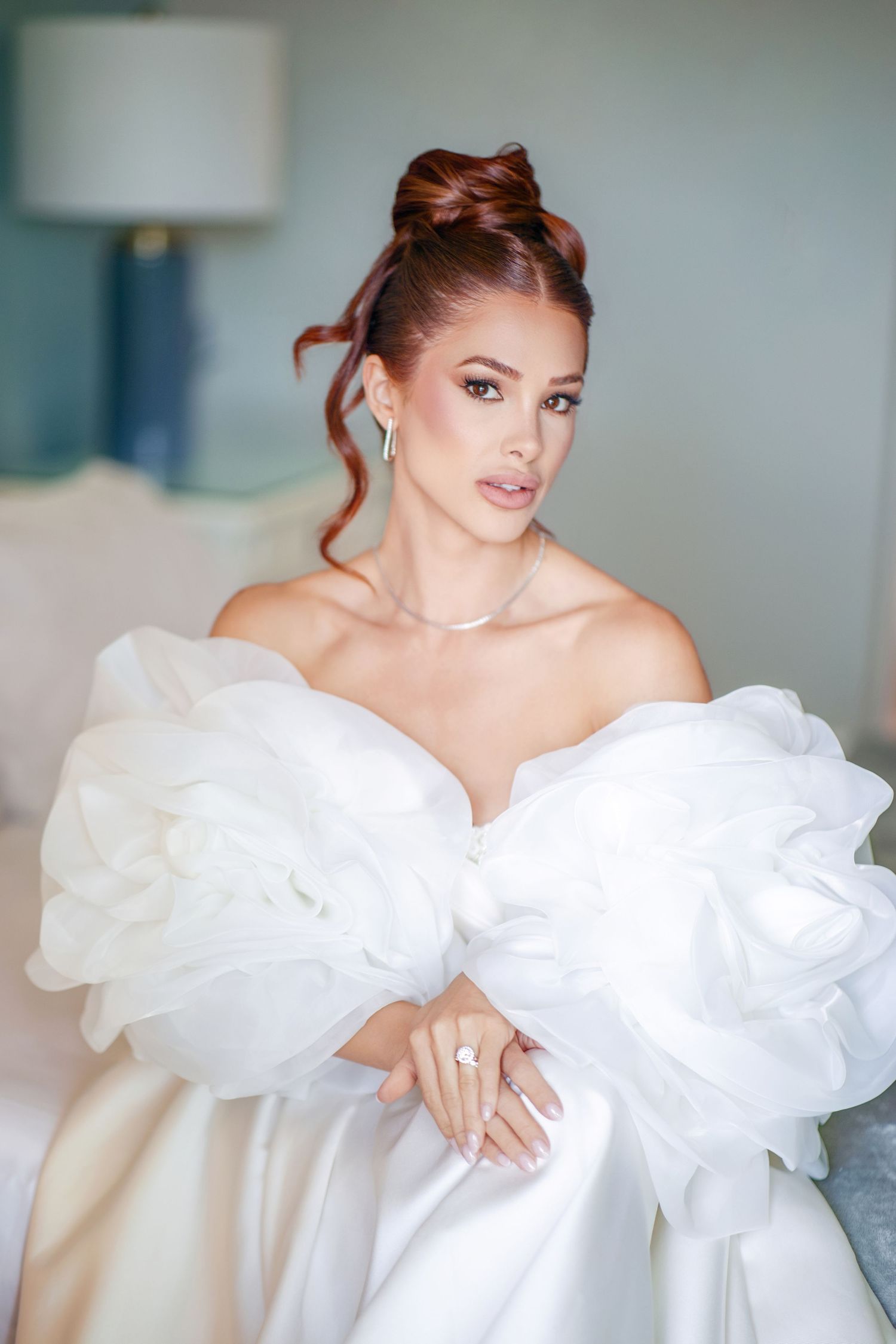 Elegant bride in white off-shoulder gown with ruffled sleeves, updo hairstyle, diamond jewelry, posing indoors.