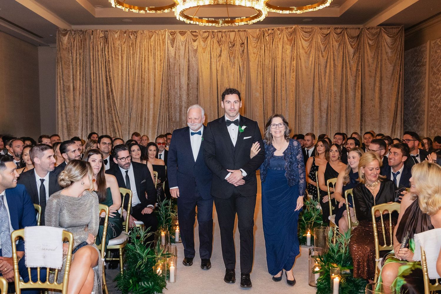 Groom walking down the aisle with his parents during an elegant indoor wedding ceremony.
