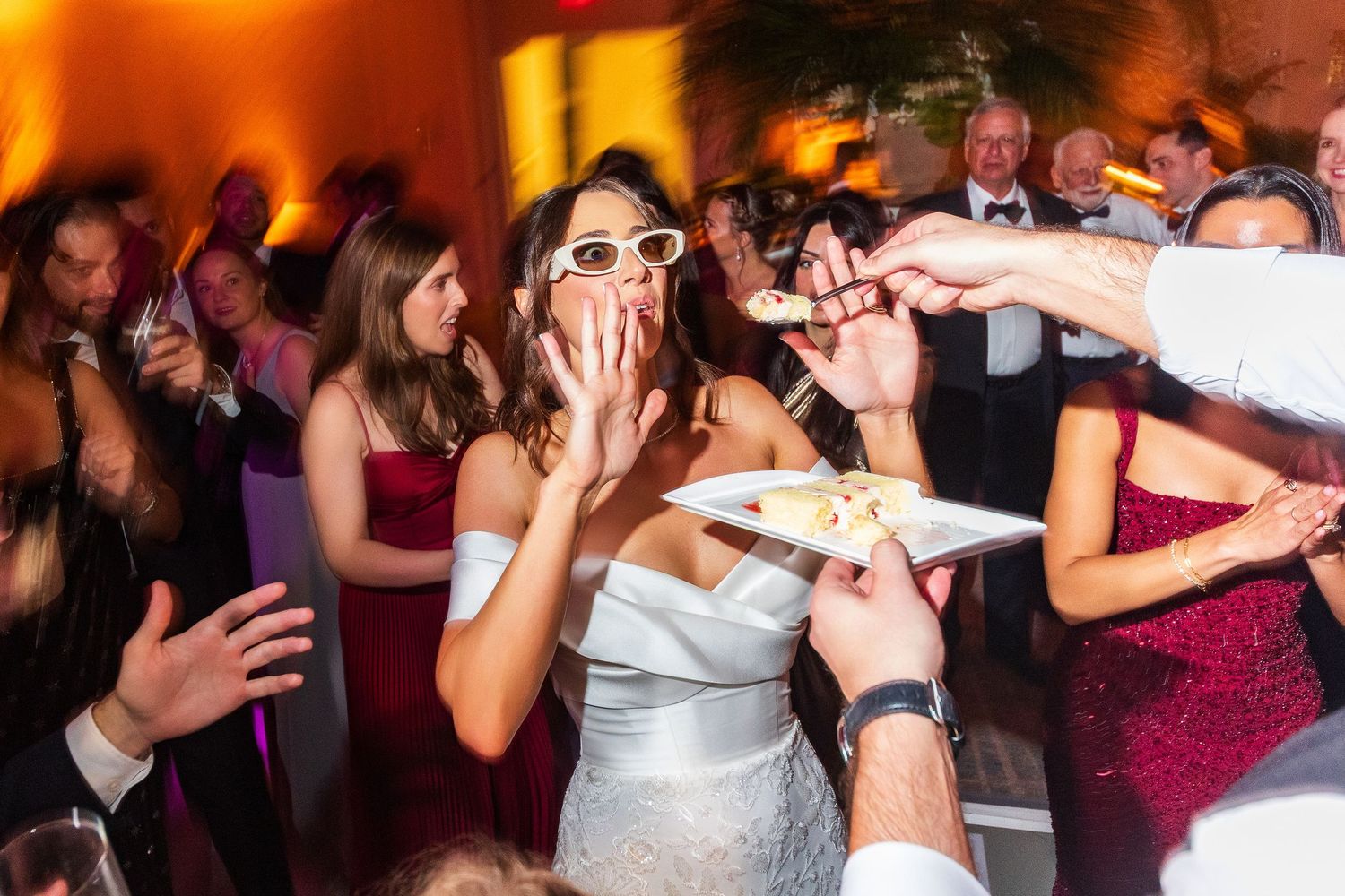 Bride in white dress and sunglasses laughing while being served wedding cake on a crowded dance floor.