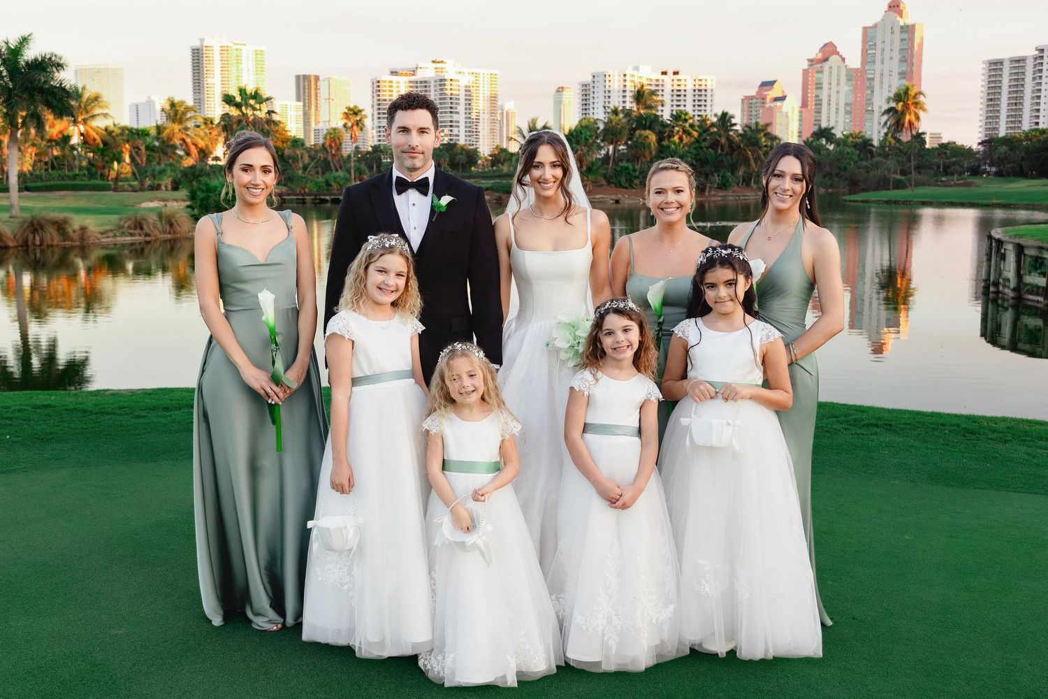 Wedding party poses outdoors with bridesmaids in sage green gowns, flower girls in white, and groom in tuxedo by a lake.